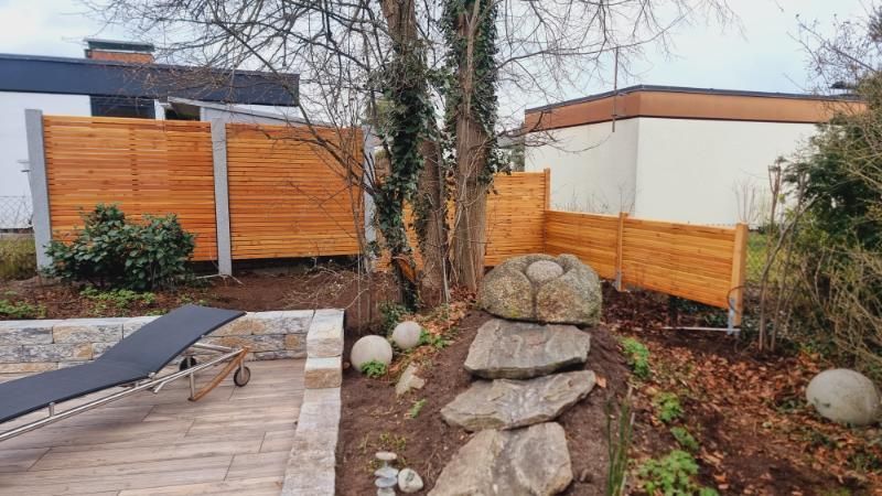 Terrasse mit grauen Pflastersteinen und einem braunen Lattenzaun neben einer ruhigen Wohnstraße