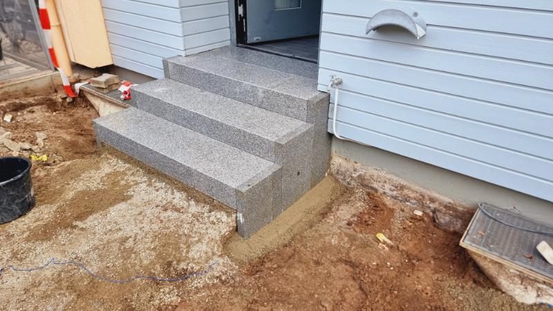 Betonierte Eingangstreppe mit kleiner Veranda neben einem weiß getünchten Haus während der Bauphase.