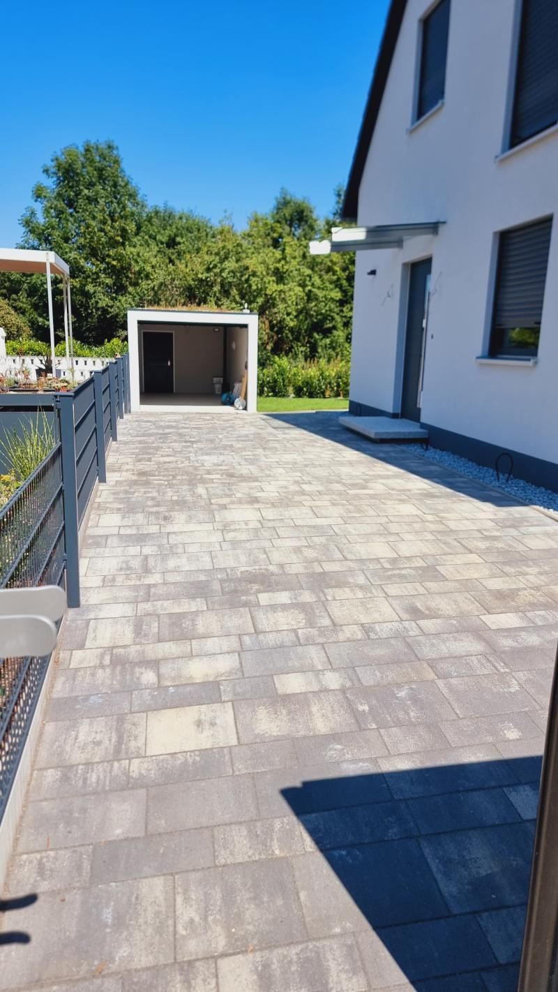 Moderne Hausterrasse mit grauen Steinplatten, einem kleinen Schuppen und Bäumen unter klarem, blauem Himmel