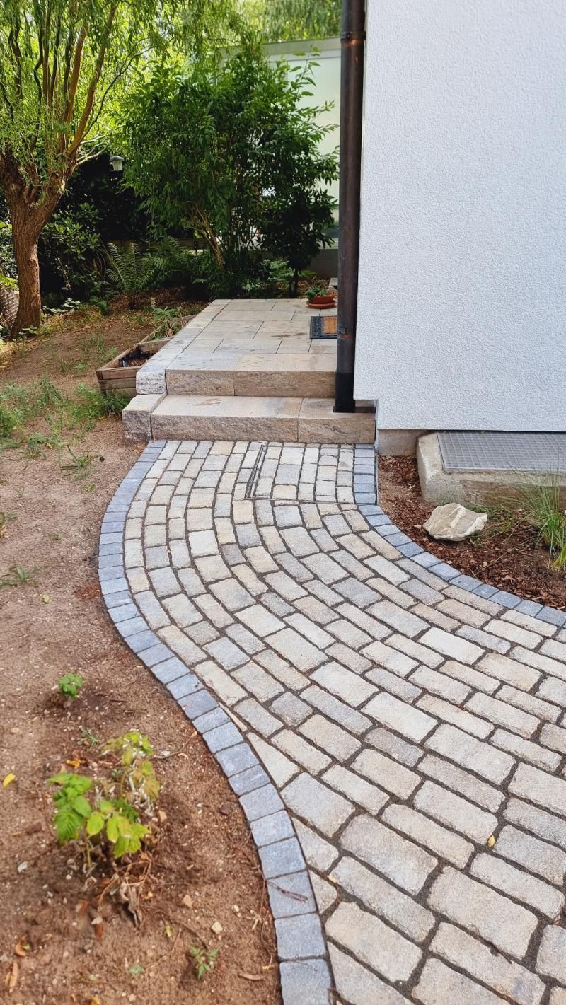 Ein geschwungener Steinweg führt neben einem weißen Gebäude durch einen Garten mit Bäumen und Sträuchern.