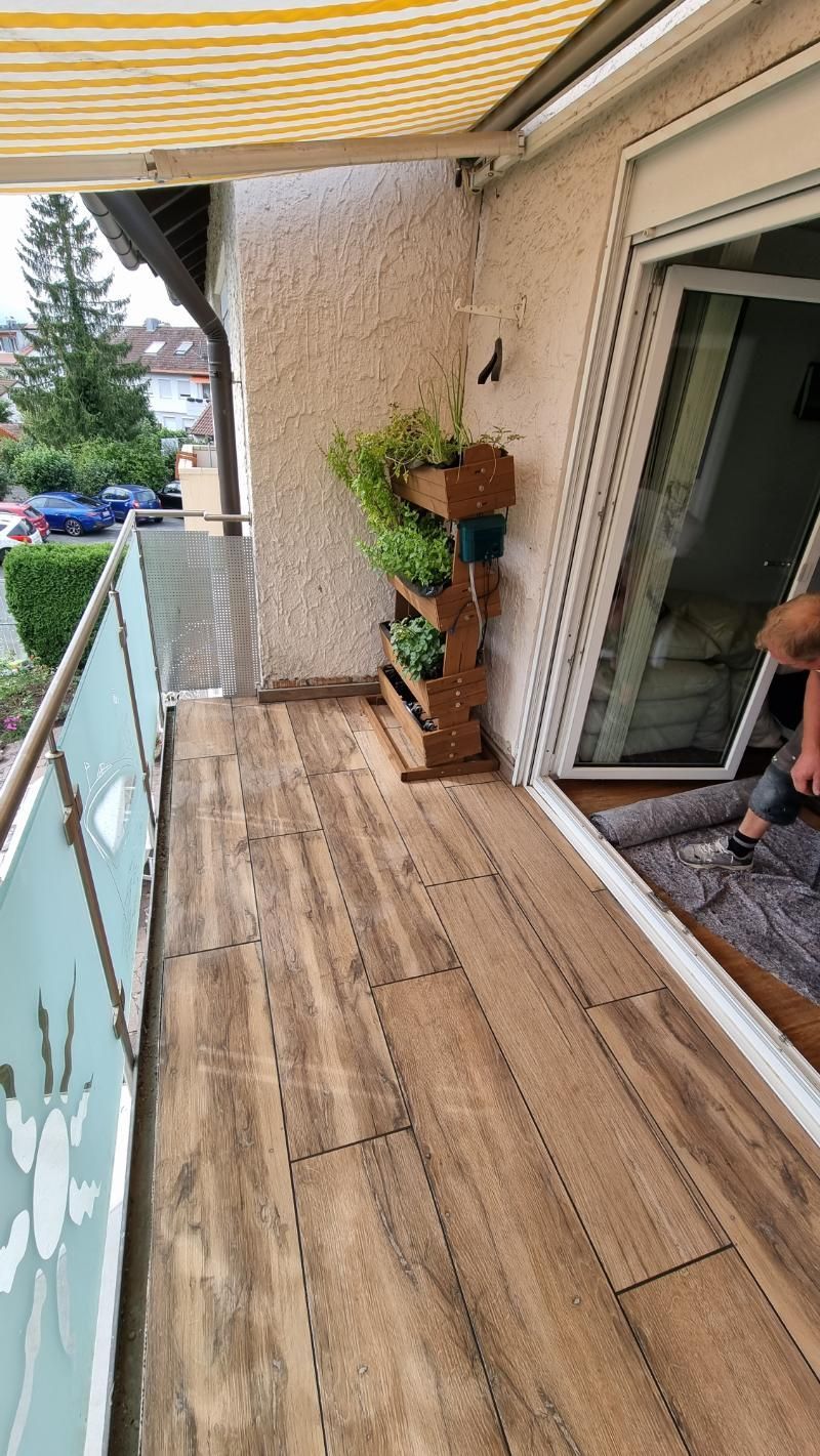 Holzbalkon der Wohnung mit Topfpflanzen, Markise darüber und einer Glasschiebetür, die sich zum Innenraum öffnet