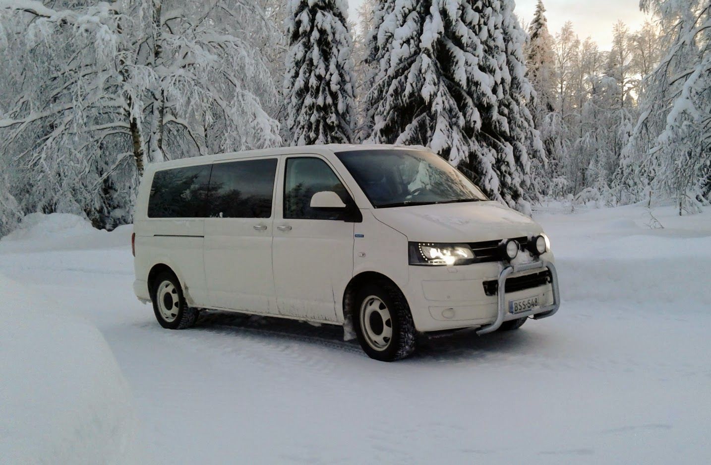 Valkoinen pakettiauto ajaa lumipeitteisellä tiellä lumisessa metsässä.