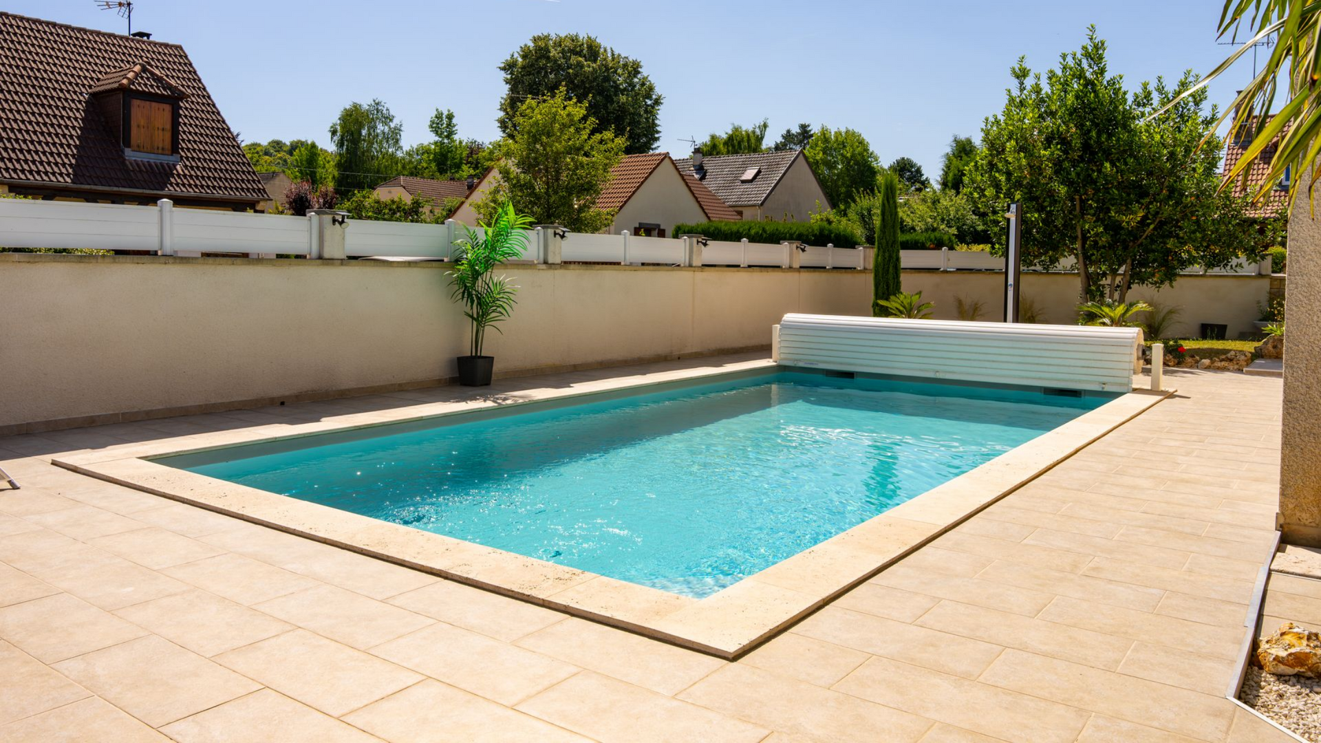 Piscine rectangulaire dans un jardin entouré de dalles beiges, d'un mur blanc et d'arbres verts.