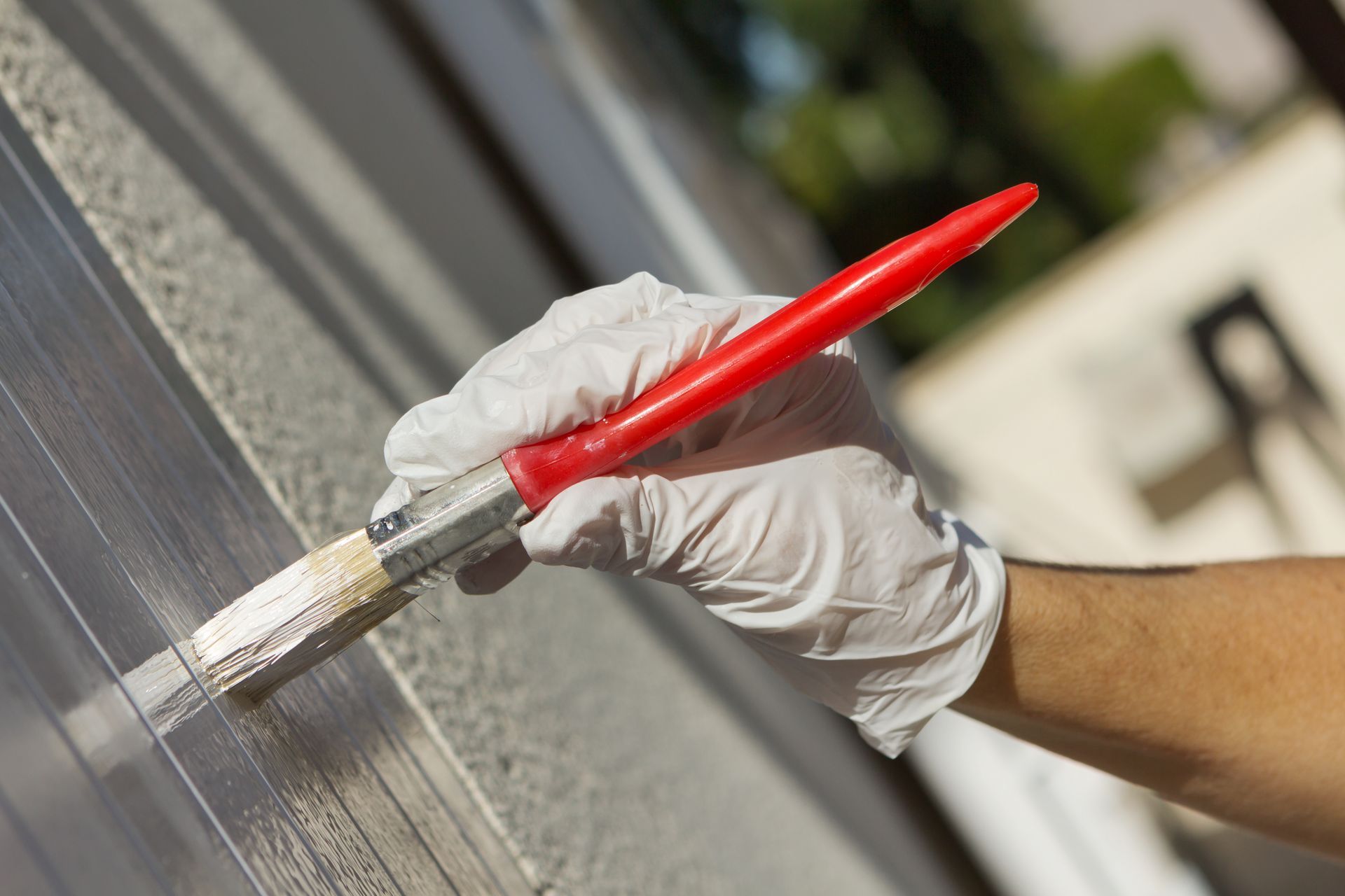 Main gantée tenant un pinceau rouge, peignant une surface claire ; en extérieur, à la lumière du jour.