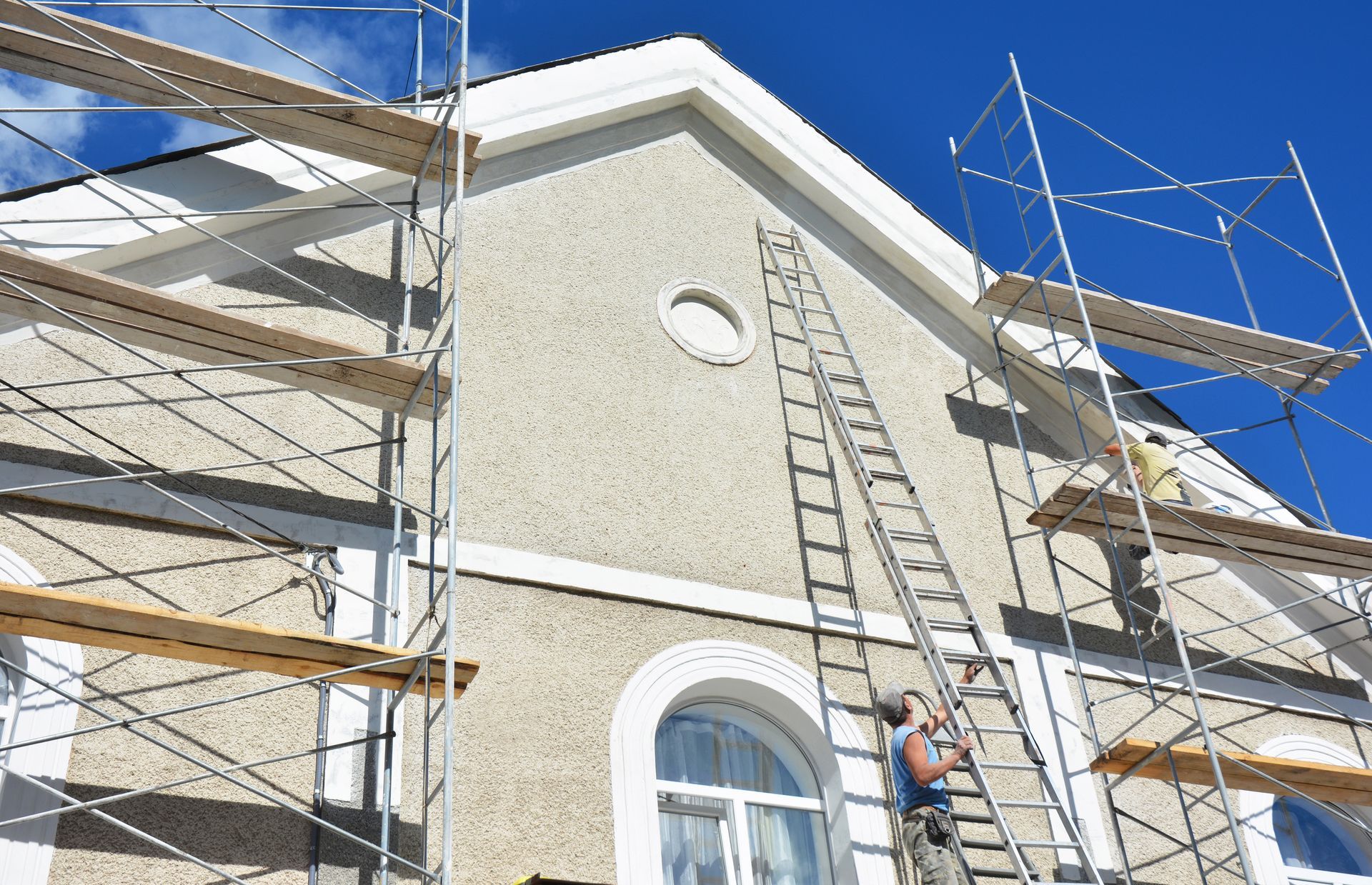 Extérieur d'un bâtiment avec échafaudage et échelle, un ouvrier peint des moulures sur fond de ciel bleu éclatant.