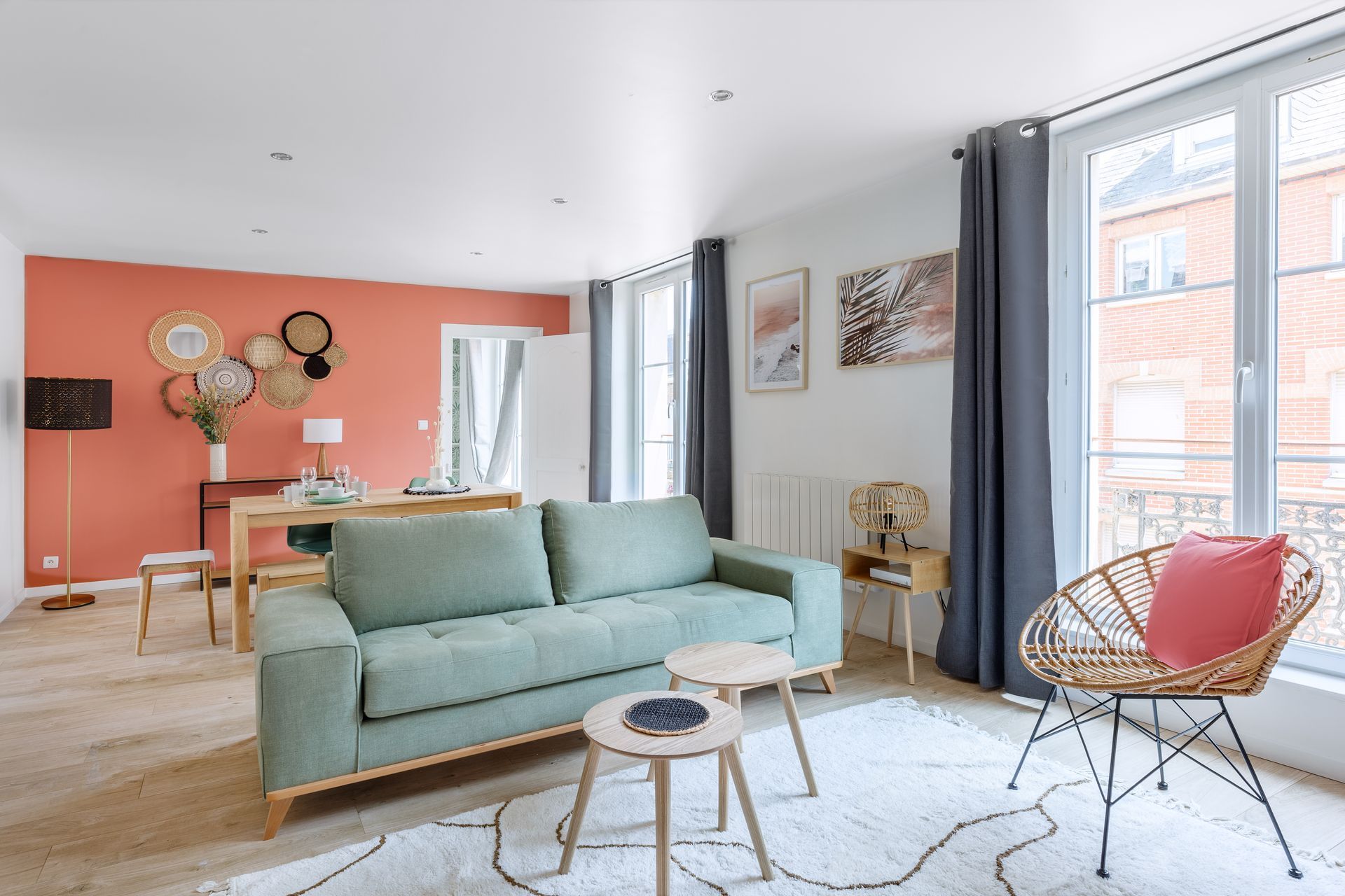 Salon avec un mur d'accent corail, un canapé vert, des meubles en bois et de grandes fenêtres avec des rideaux gris.
