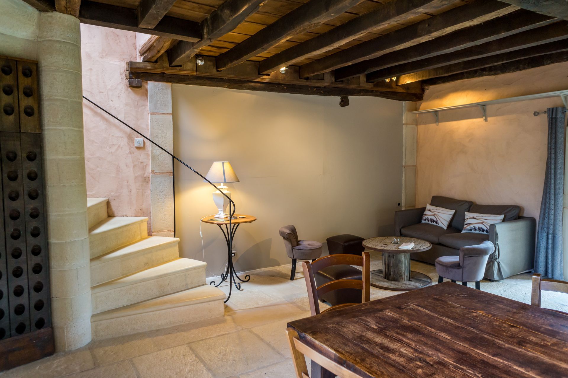 Un escalier en pierre mène à un espace de vie rustique avec poutres apparentes, canapé, chaises et table.
