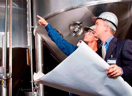 Dos hombres con cascos de seguridad examinan planos junto a tanques industriales.