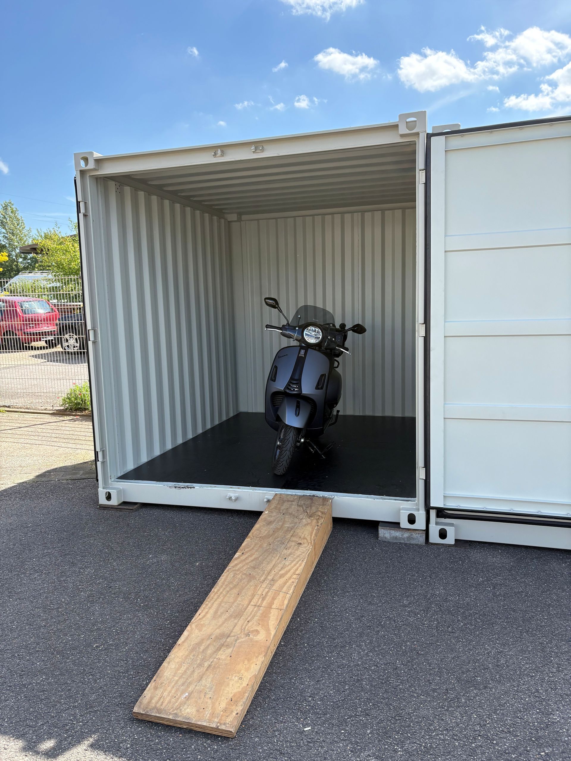 Motorrad in einem offenen weißen Schiffscontainer mit einer Holzrampe, die zu Asphalt hinunterführt.