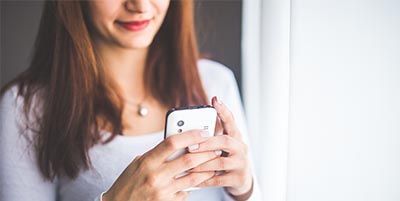 Eine Frau hält vor einem Fenster ein Mobiltelefon in ihren Händen.