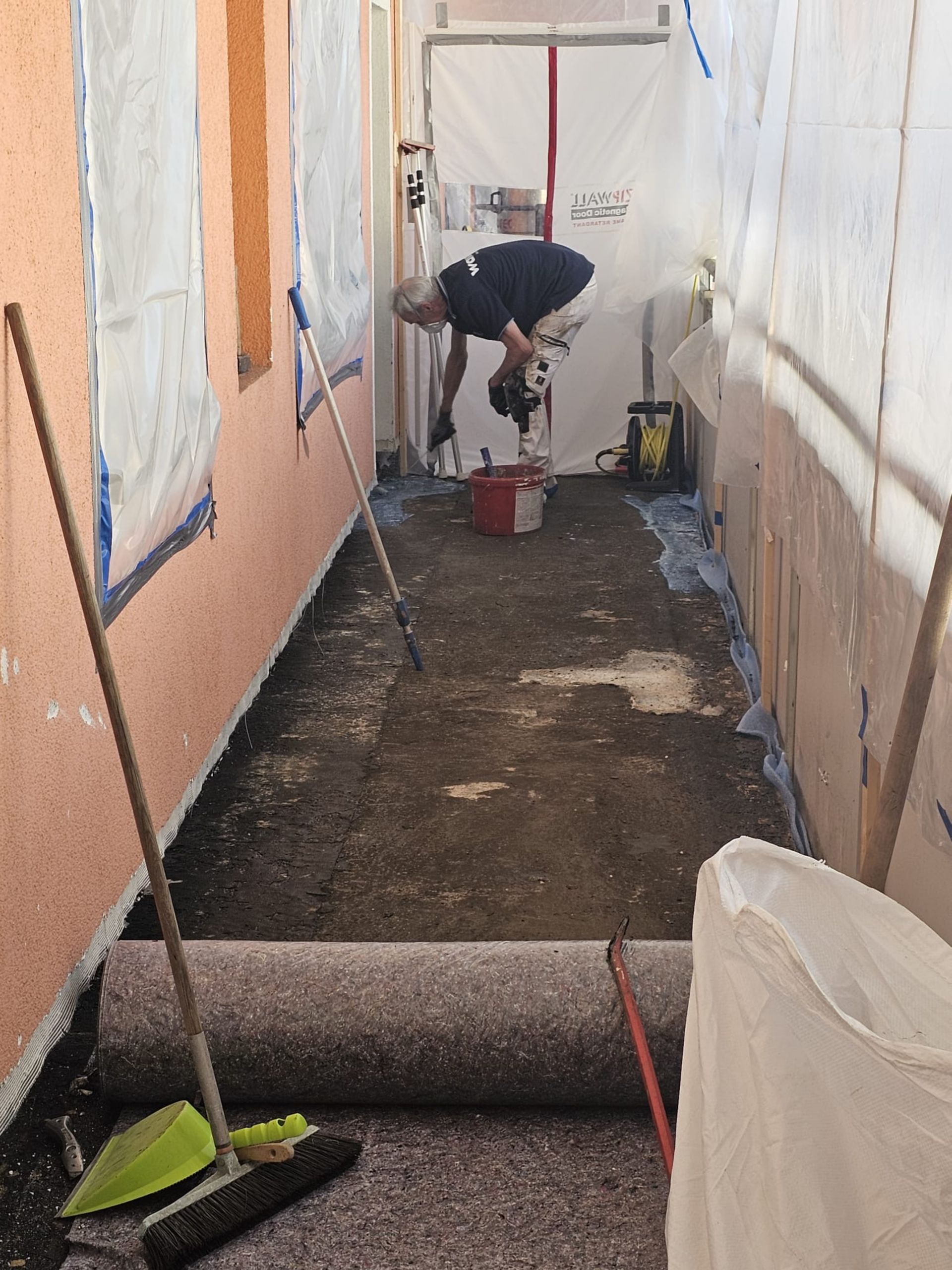 Ein Arbeiter streicht einen schmalen Außendurchgang, dessen Wände abgeklebt sind; eine Farbrolle steht auf dem Boden.