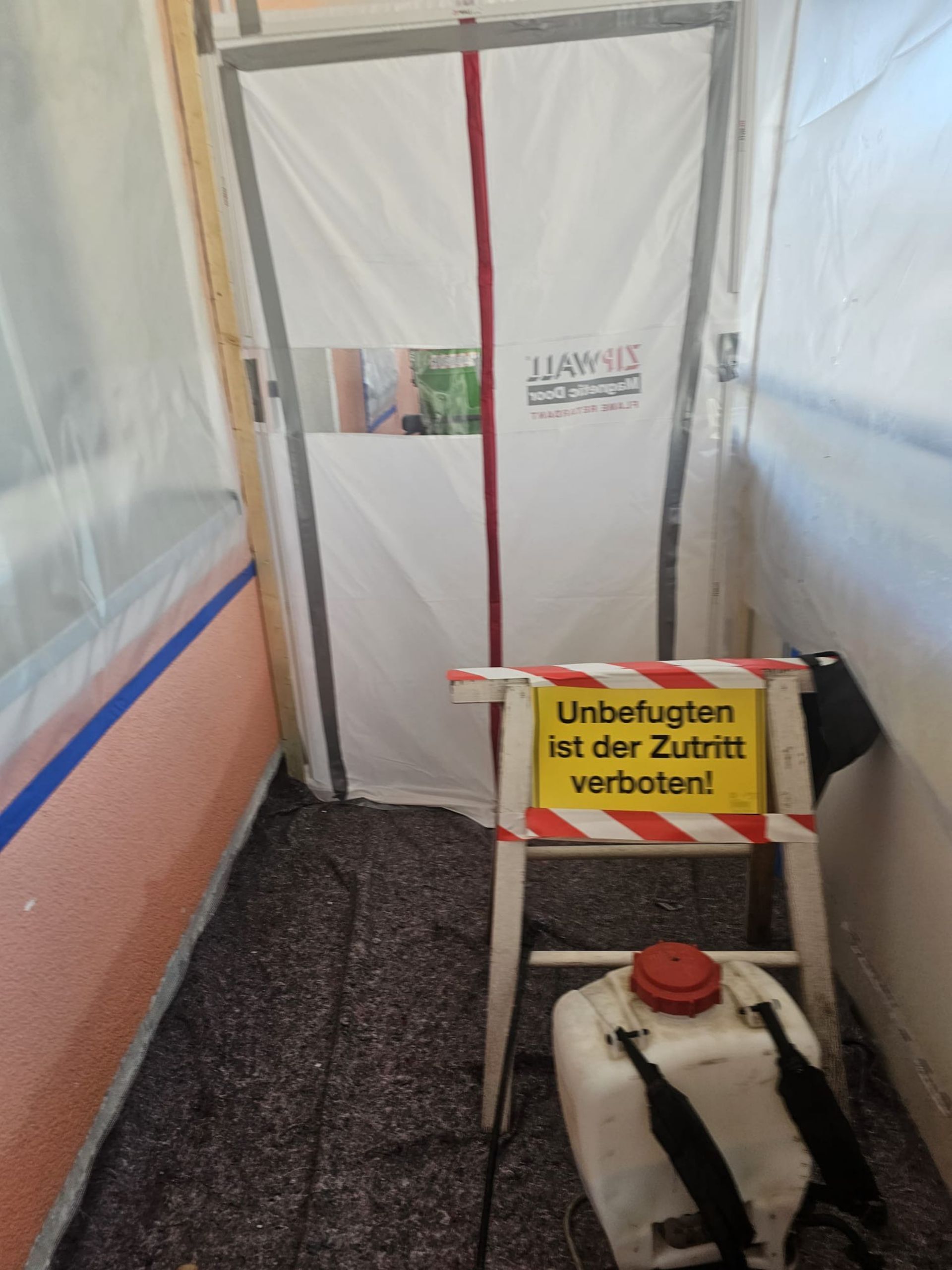 Warnschild auf einer kleinen grauen Leiter in einem mit Plastikfolie abgedeckten Flur, daneben ein roter Benzinkanister.