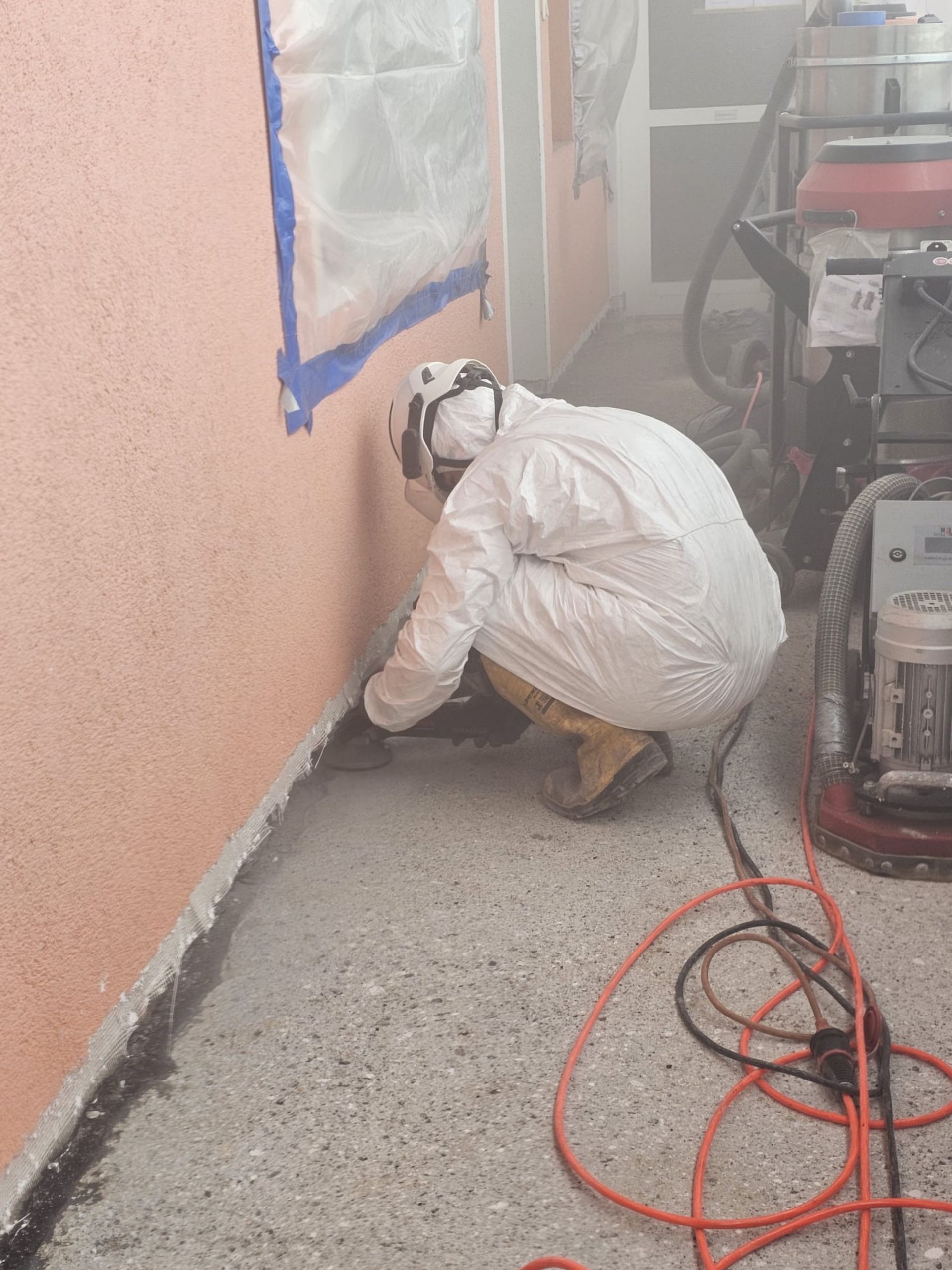 Ein Arbeiter in weißem Schutzanzug kniet neben einer Wand, deren Ränder blau besprüht sind; auf dem Boden liegen Kabel.
