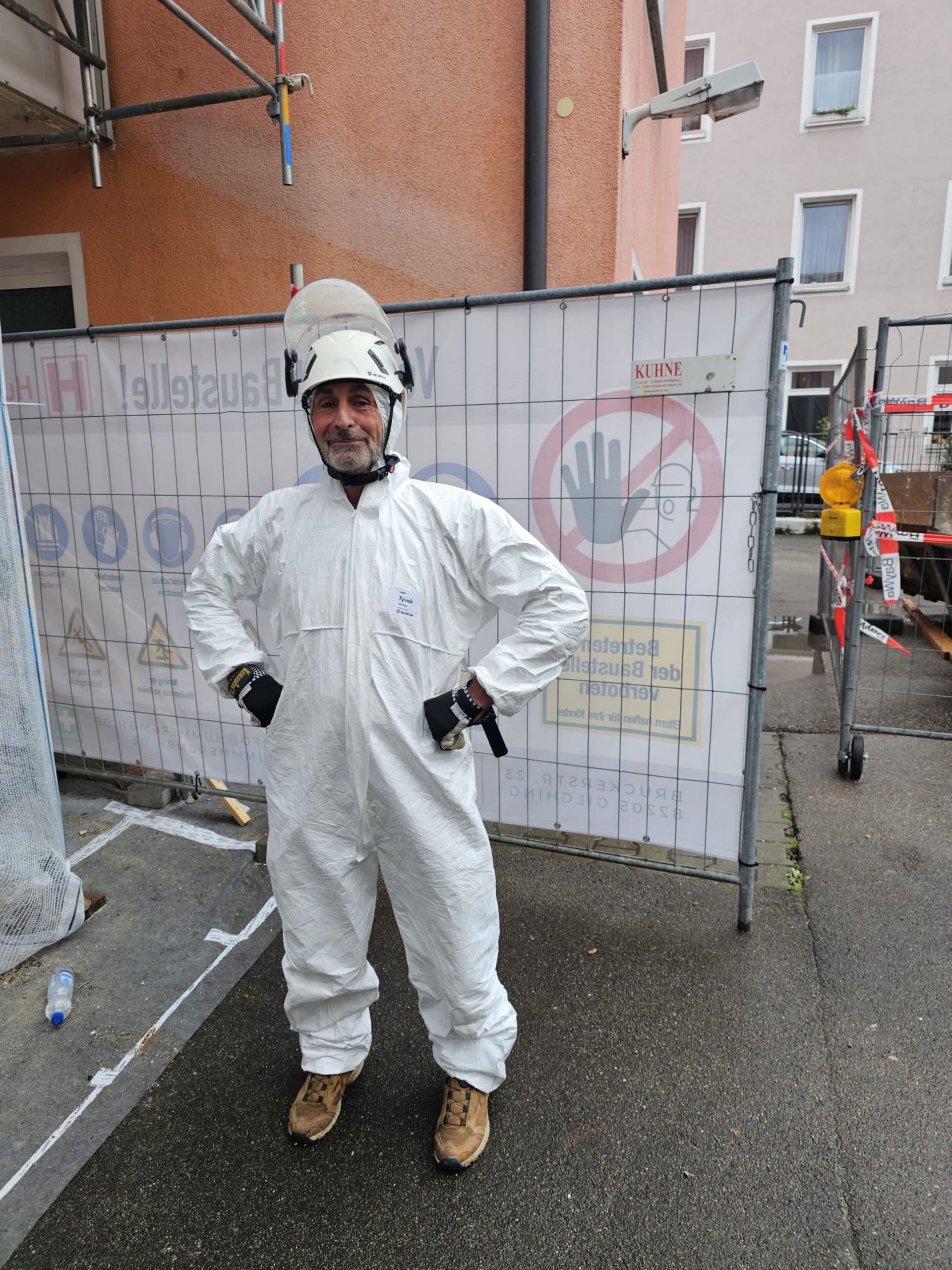 Ein Arbeiter in weißem Schutzanzug und Helm steht an einem Baustellenzaun.