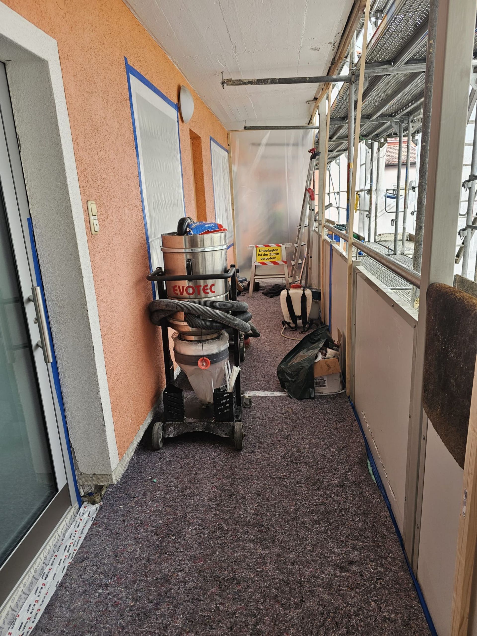 Schmaler Außengang mit Bauwerkzeugen, Plastikplanen und Bauschutt entlang einer Gebäudewand.