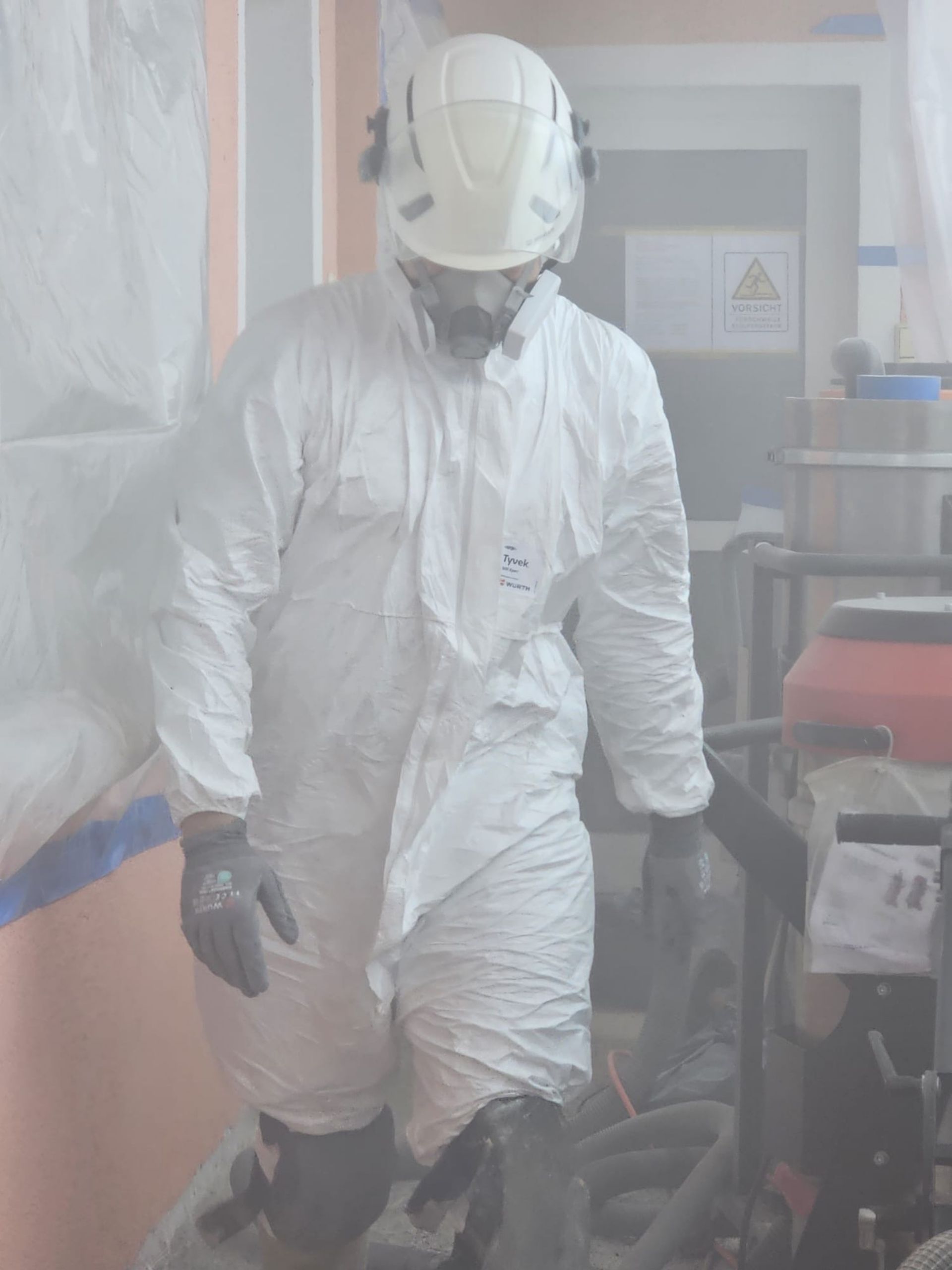 Eine Person in weißem Schutzanzug und Helm geht durch ein diesiges Industriegebiet mit Ausrüstung und Containern.