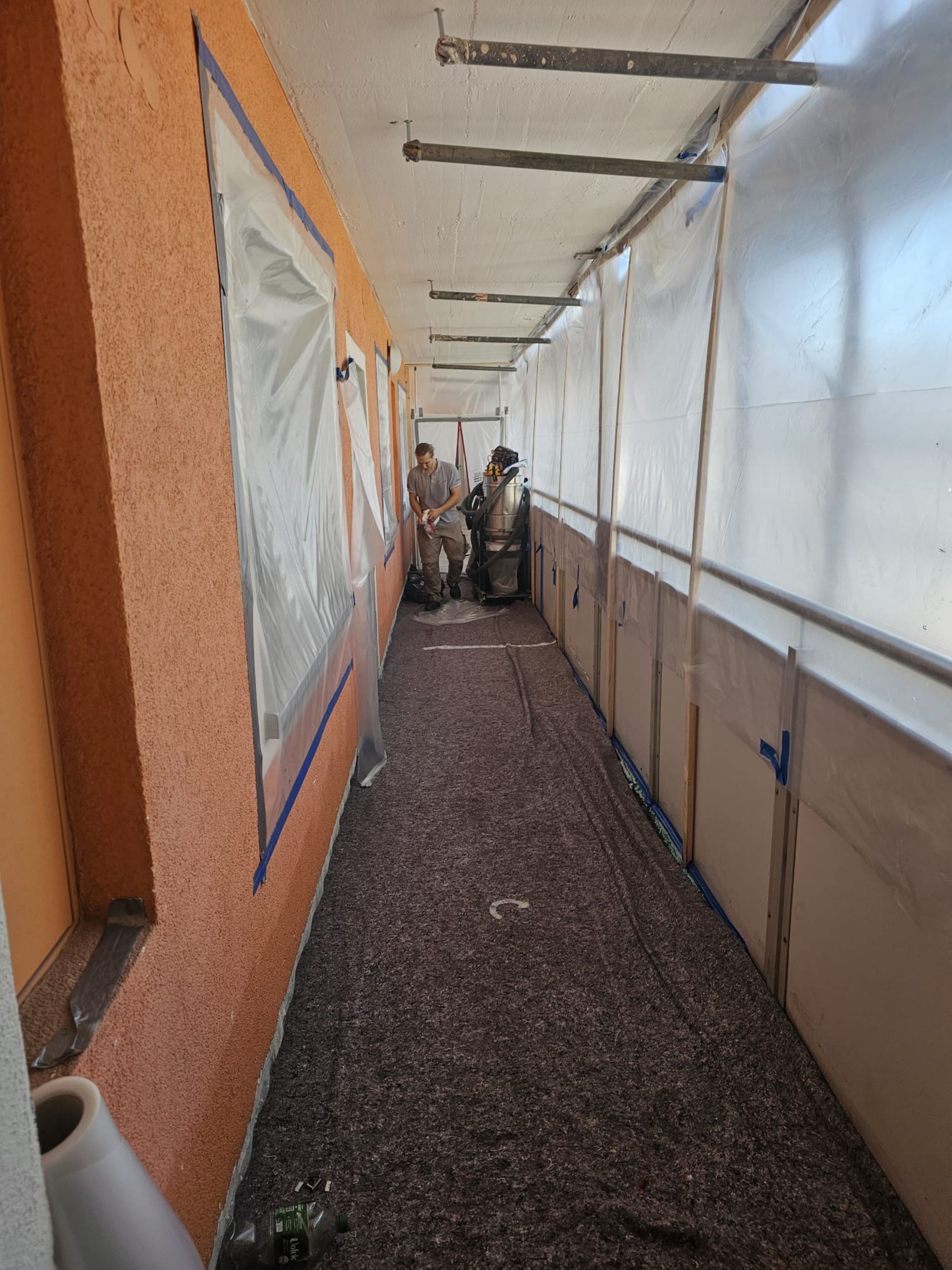 Schmaler Außengang mit orangefarbener Wand, weißen Planen, Kiesboden und Menschen am anderen Ende