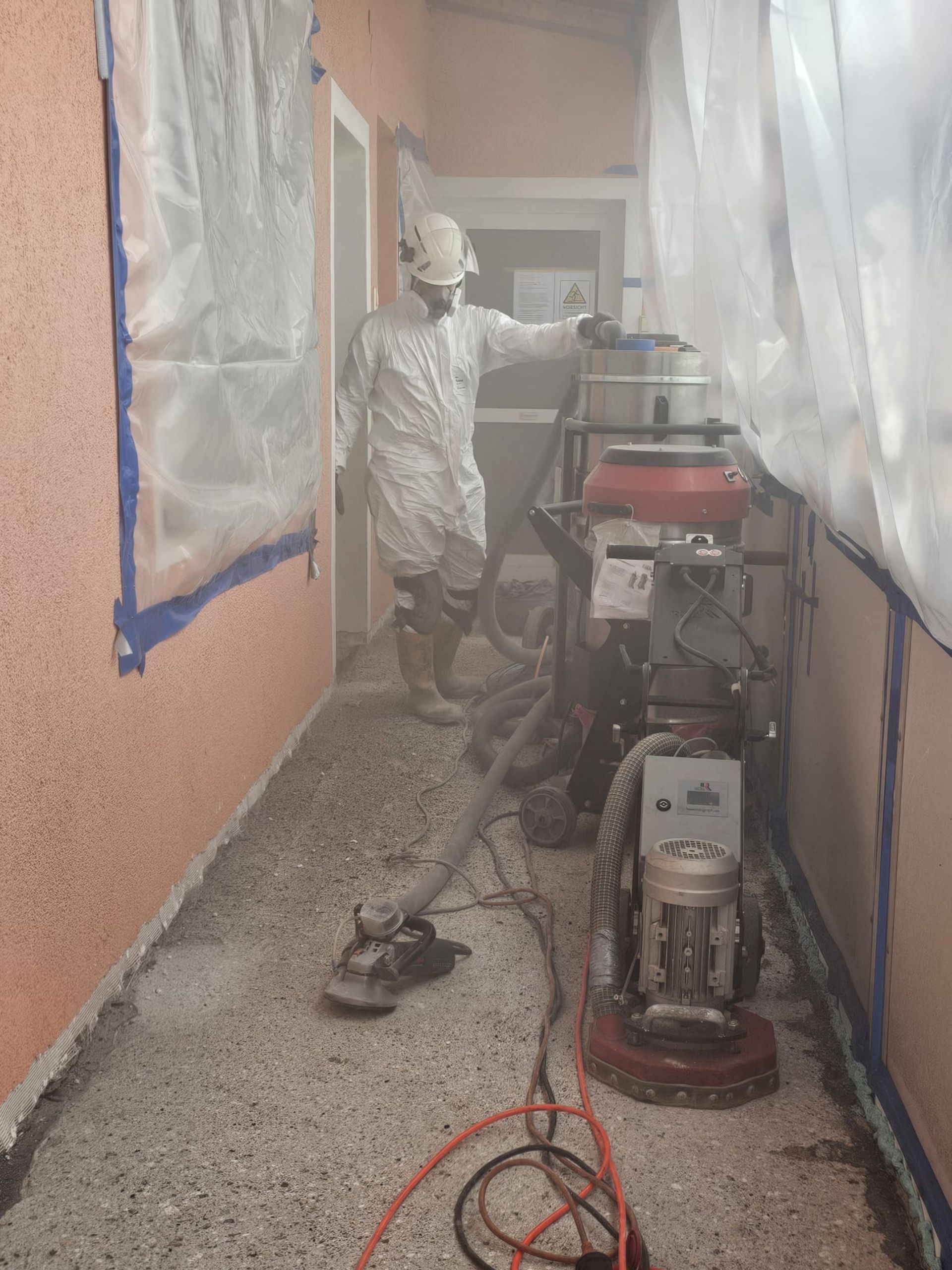 Ein Arbeiter in Schutzkleidung besprüht im Rahmen einer Oberflächenbehandlung einen schmalen Korridor mit Geräten und Schläuchen.