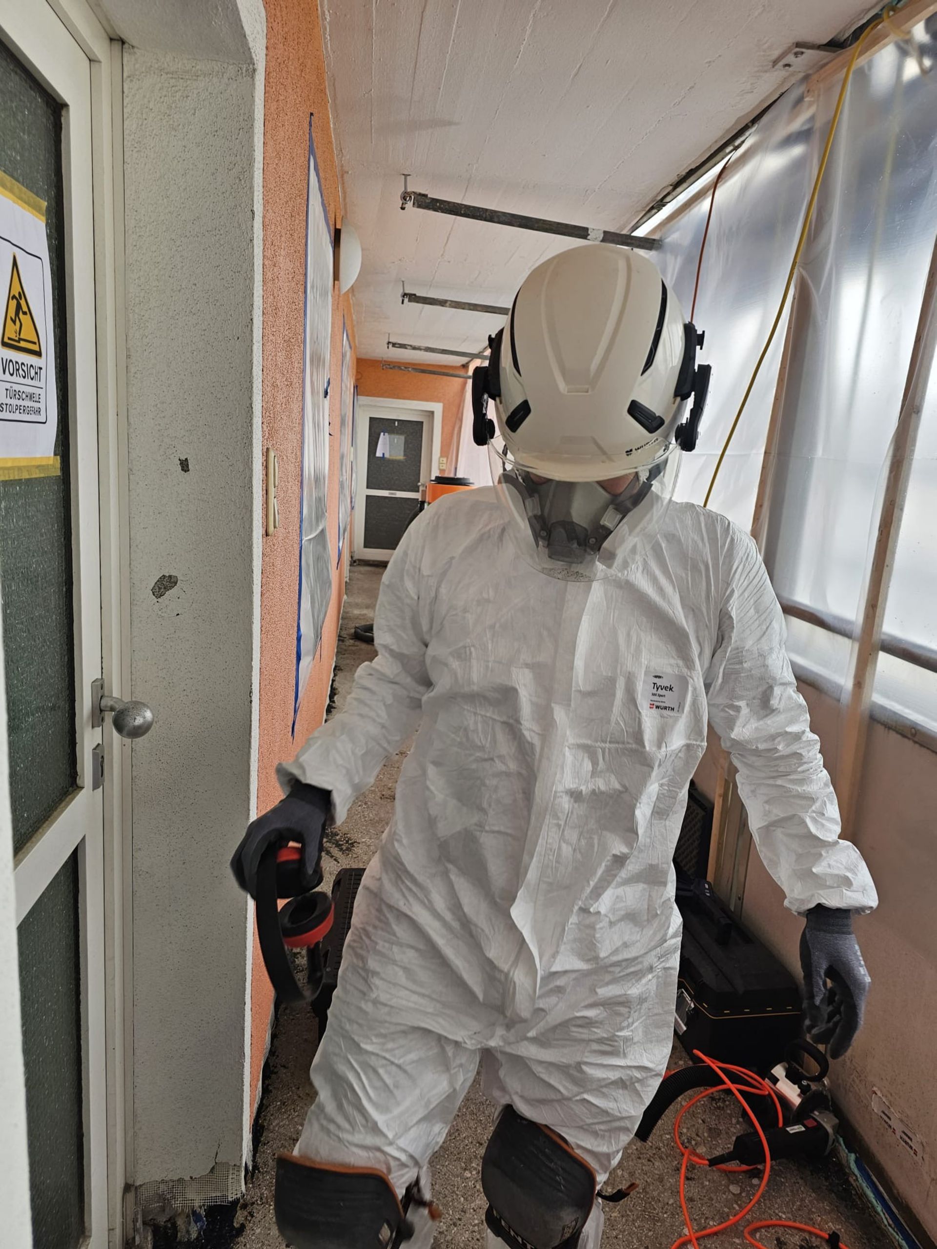 Eine Person in weißem Schutzanzug und Helm steht in einem schmalen Flur und hält Ausrüstung und Kabel in der Hand.