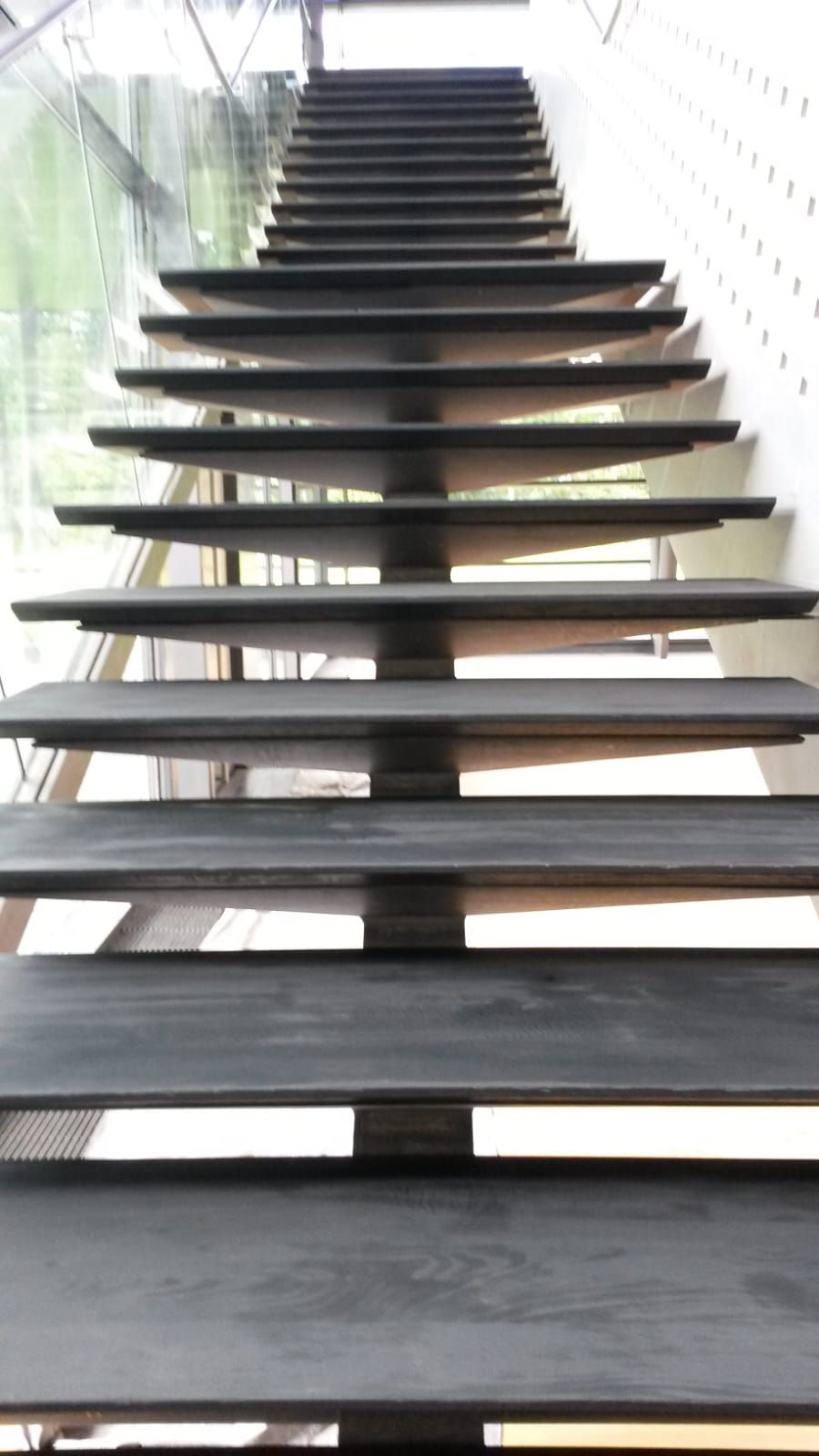 Treppe mit dunklen Metallstufen, die nach oben führt. Auf der linken Seite befindet sich ein Glasfenster, auf der rechten Seite eine weiß gesprenkelte Wand.