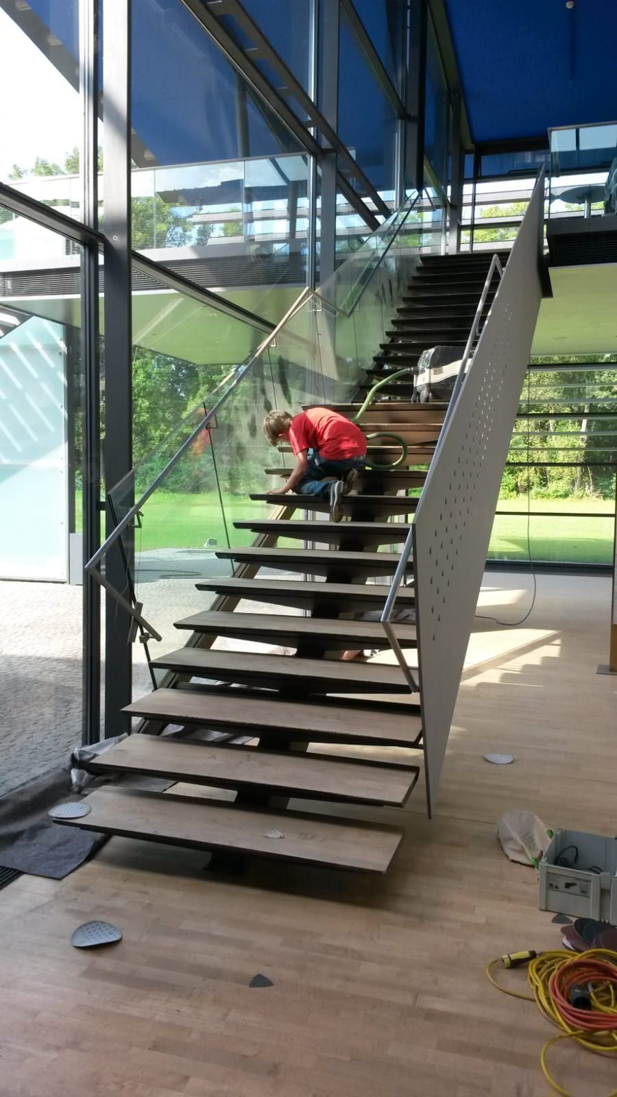 Person im roten Hemd arbeitet an einer modernen Treppe aus Stahl und Holz in einem Gebäude mit Glaswänden.