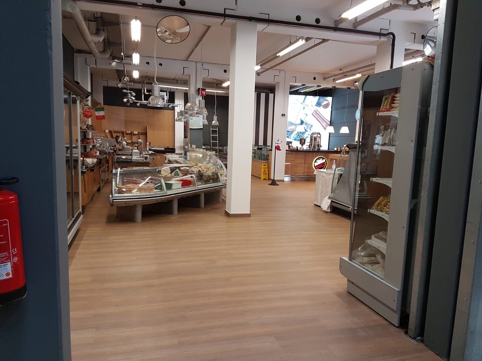 Innenansicht eines Feinkostladens: Holzboden, Vitrinen mit Lebensmitteln, weiße Säulen, ein Feuerlöscher.