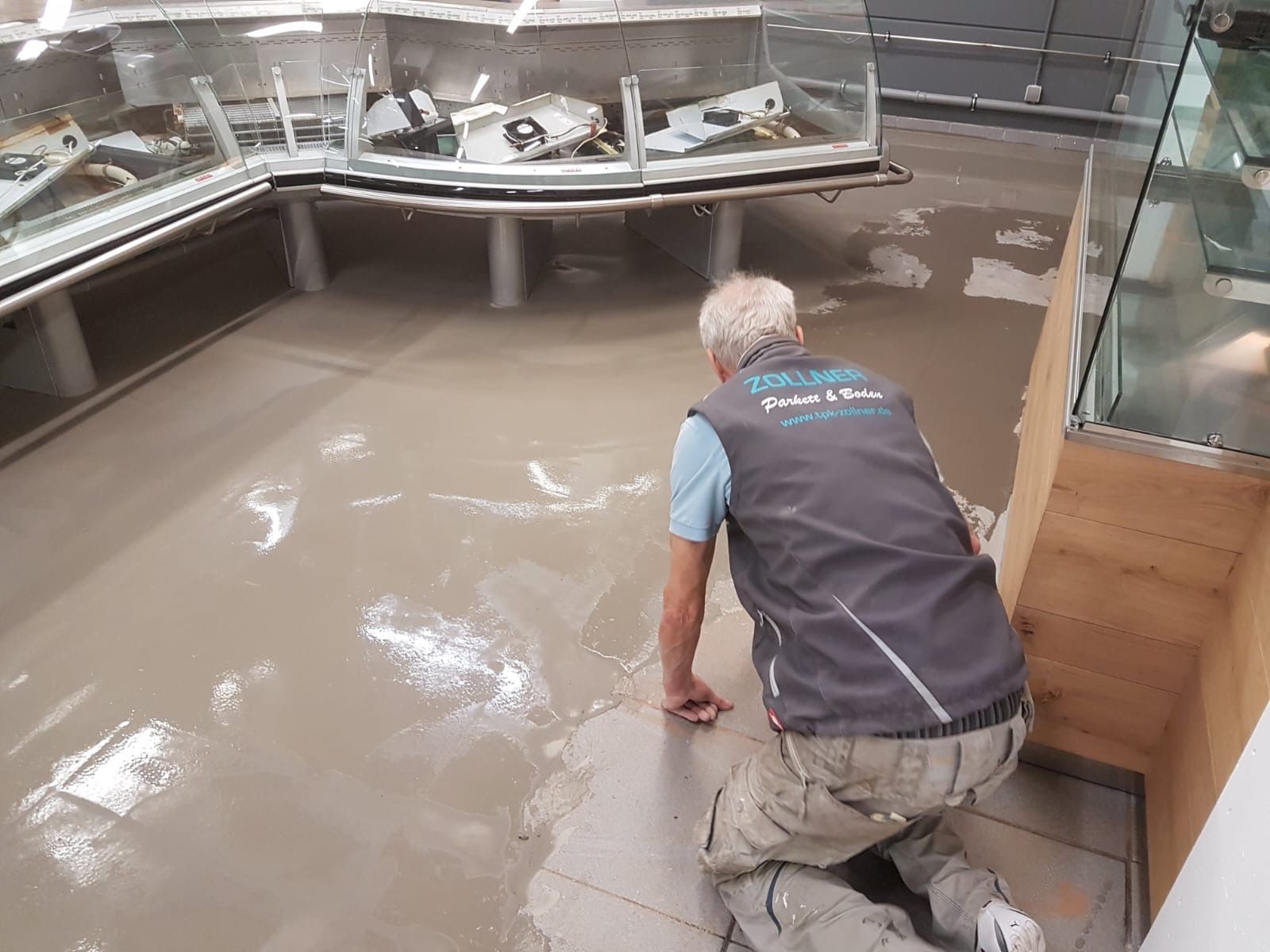 Ein Mann kniet in einem überfluteten Geschäft und betrachtet den Wasserschaden. Die Vitrinen stehen mitten im trüben Wasser.