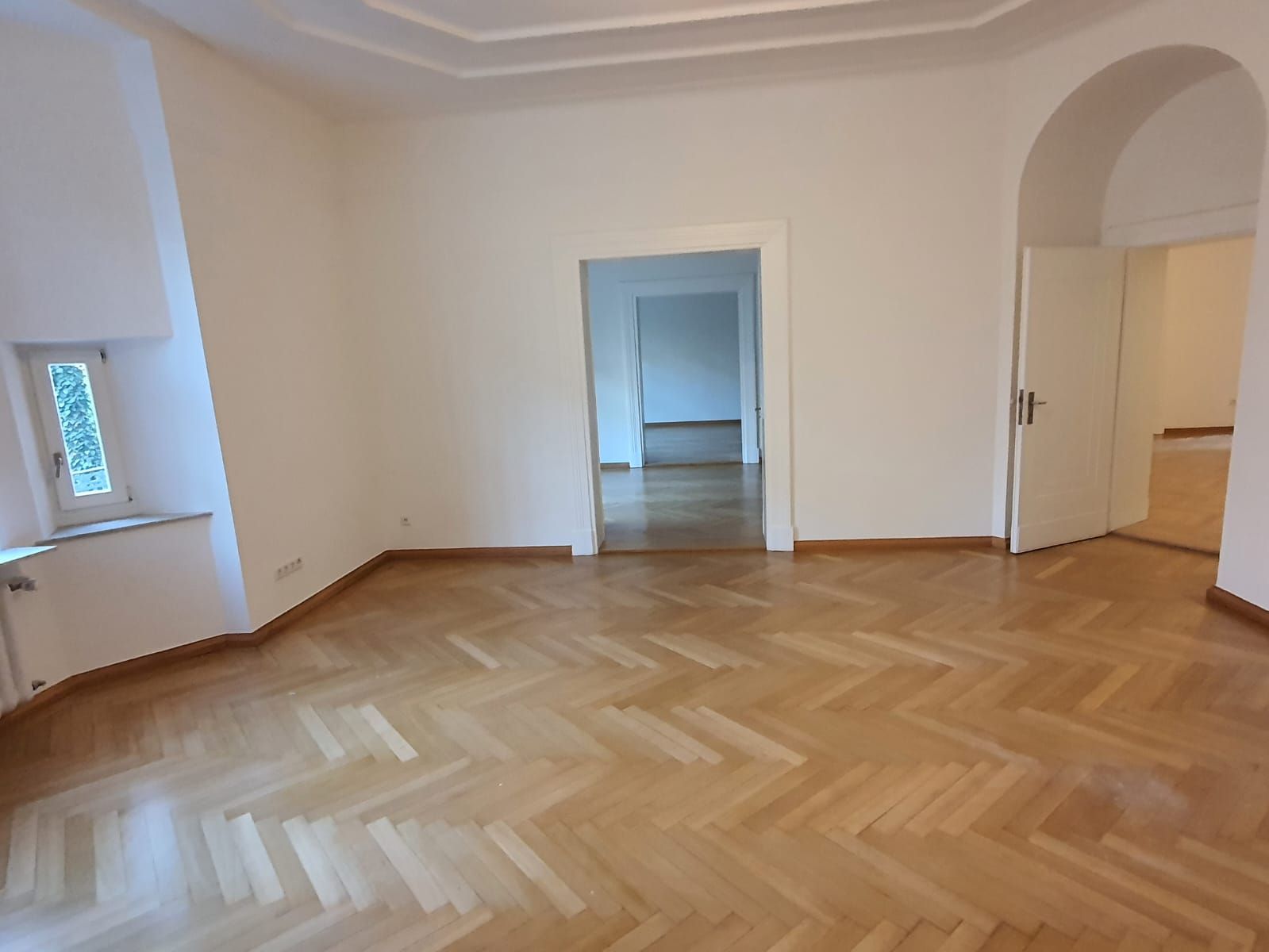 Leerer Raum mit Holzboden im Fischgrätenmuster, weißen Wänden und Türen, die zu anderen Räumen führen.