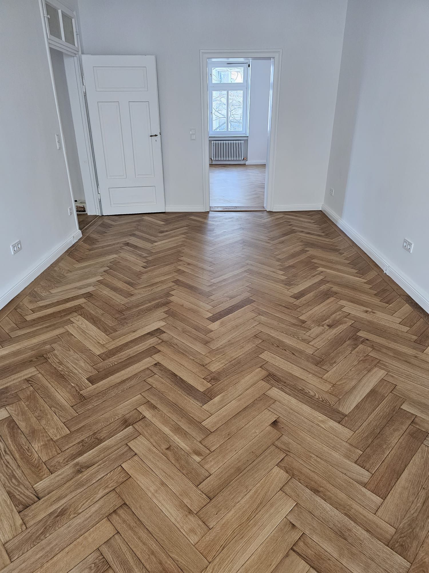 Fischgrät-Hartholzboden in einem hellen, leeren Raum. Offene Tür zu einem Flur mit sichtbarem Heizkörper.