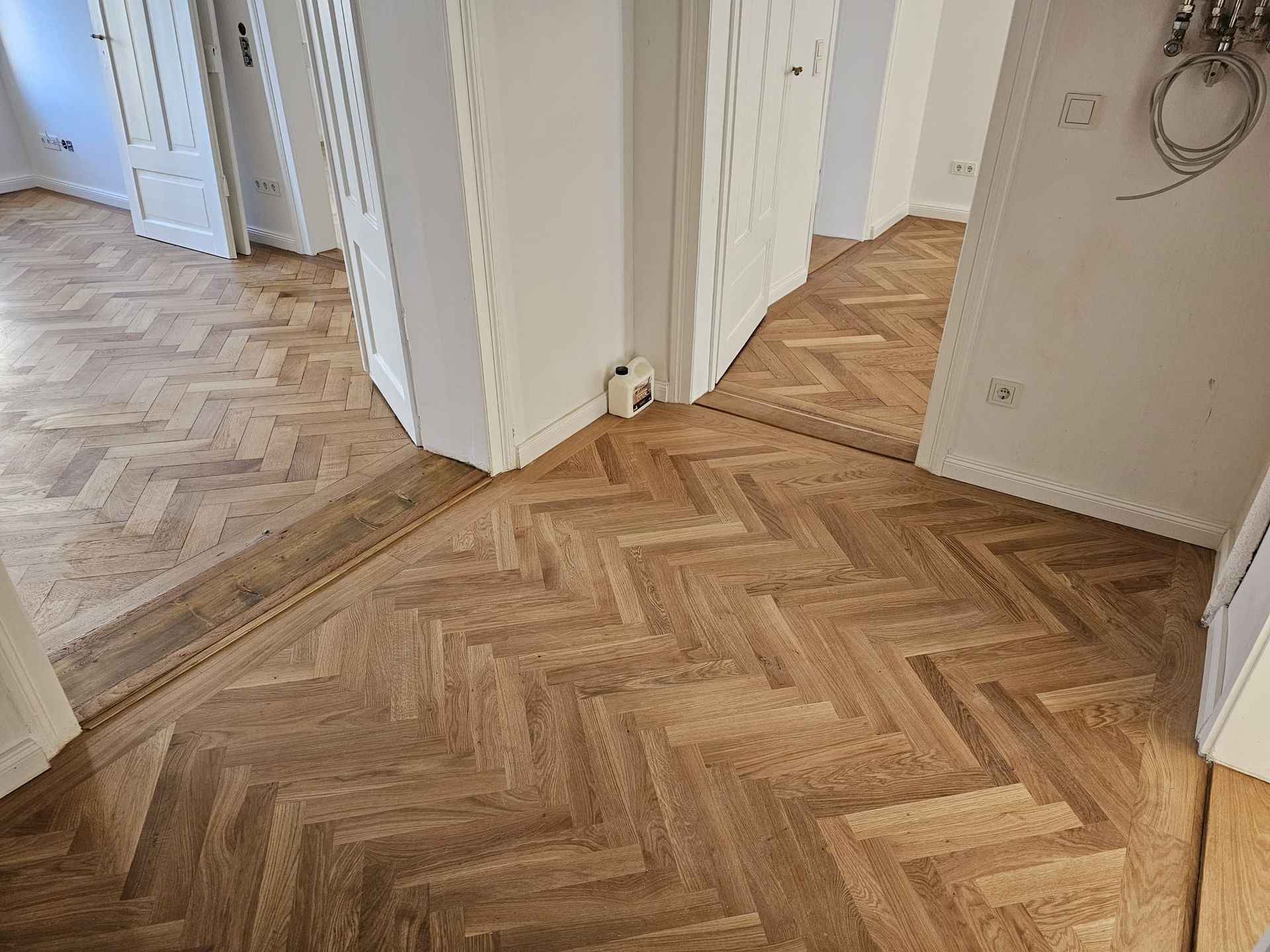 Fischgrät-Holzboden in einem Flur, eingefasst von dunklerem Holz. Weiße Wände.