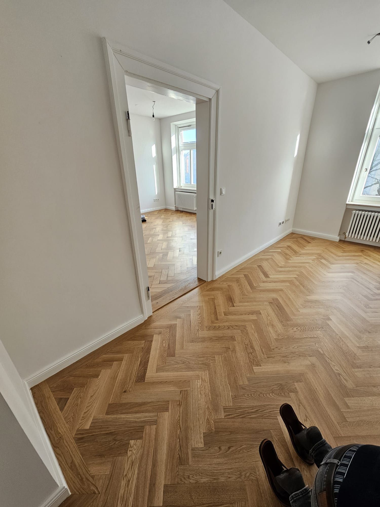 Ein Zimmer mit Fischgrätholzboden, Tür zu einem anderen Zimmer mit Fenster und weißen Wänden.