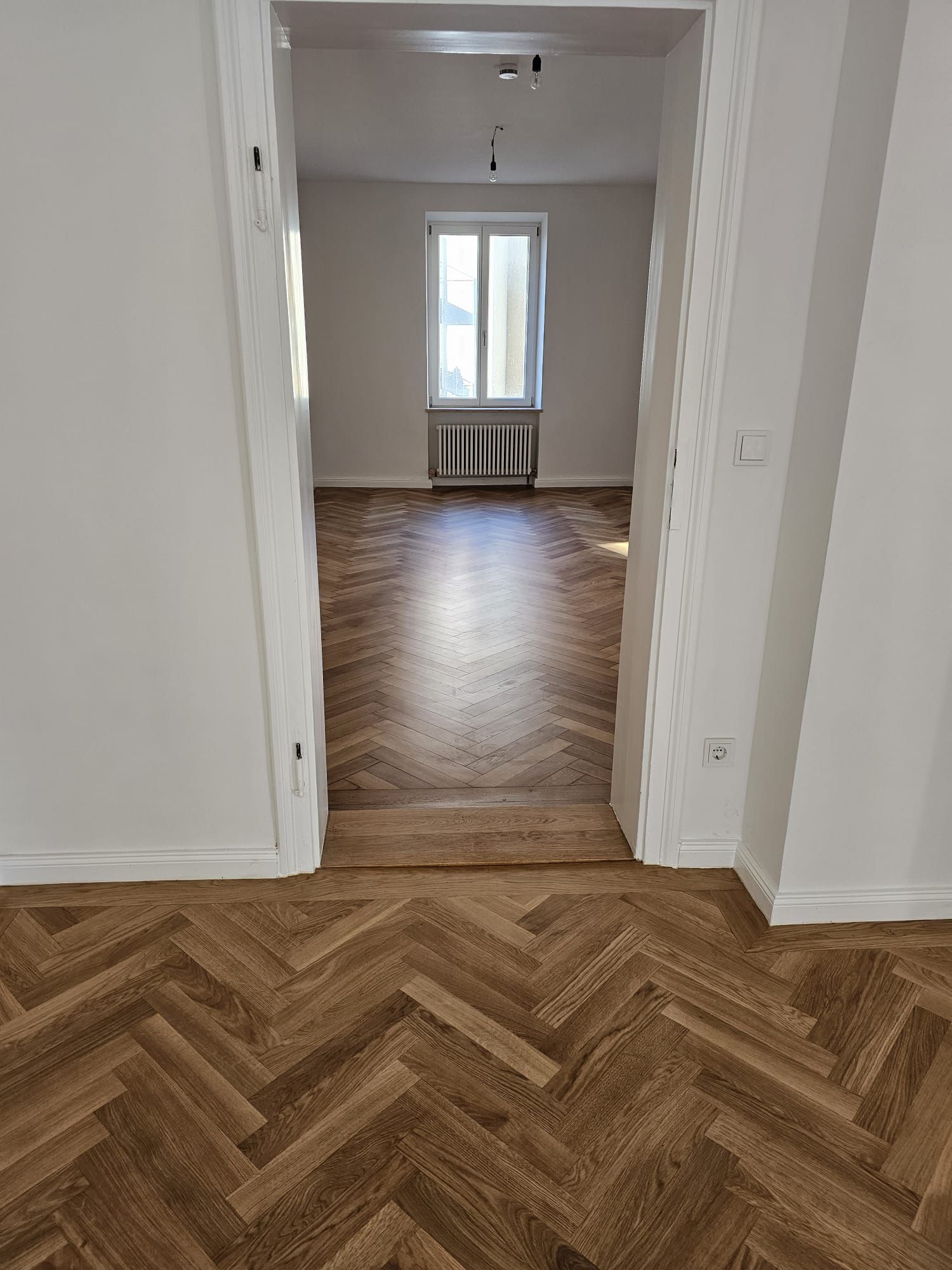 Blick durch eine offene Tür: Fischgrätholzböden führen zu einem sonnendurchfluteten Raum mit einem Fenster.