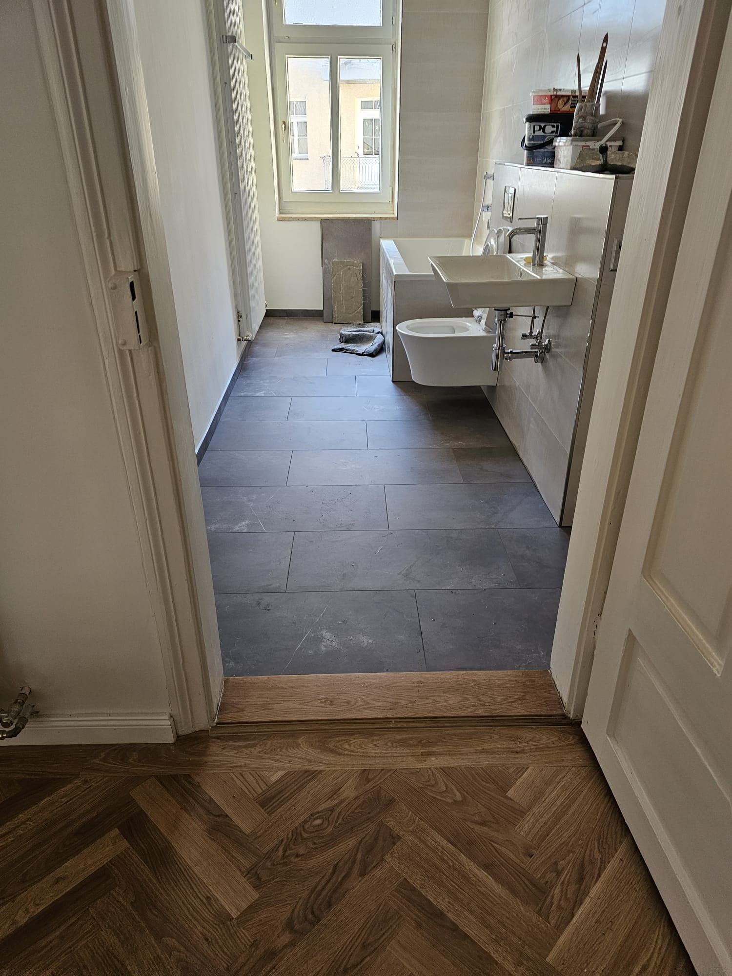 Badezimmer mit neuen grauen Bodenfliesen, offene Tür mit Blick auf den Holzboden im Flur.