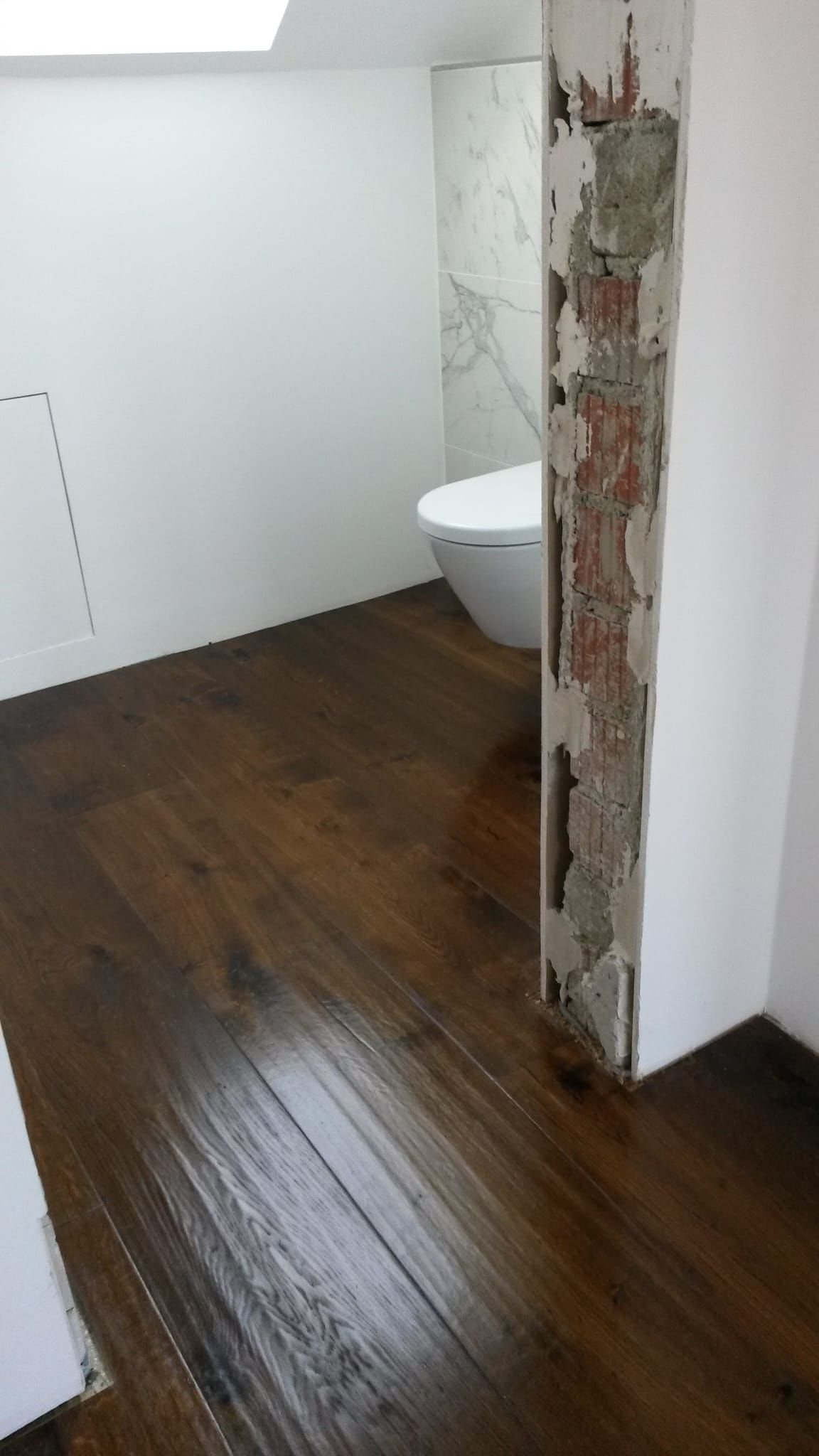 Ein dunkler Holzboden führt in ein Badezimmer mit weißer Toilette, freiliegenden Ziegelsäulen und weißen Wänden.