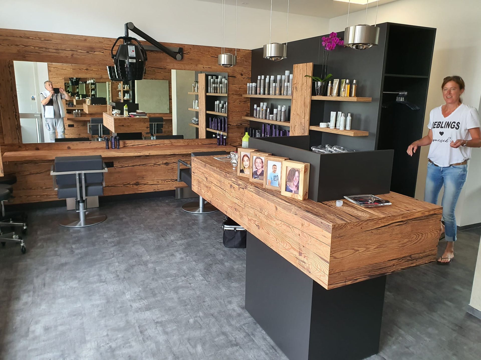 Innenansicht eines Friseursalons: Frau steht neben einem hölzernen Empfangstresen, holzgetäfelten Styling-Stationen und einer Produktausstellung.