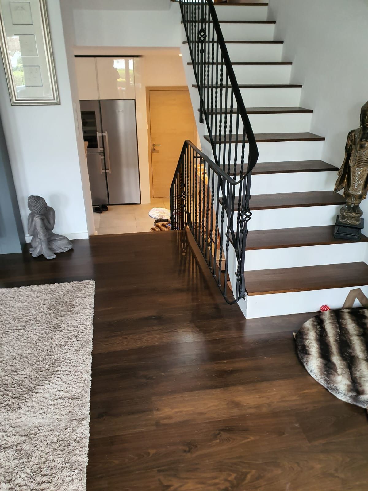 Holztreppe mit schwarzem Geländer. Ein Kühlschrank und ein Flur sind sichtbar.