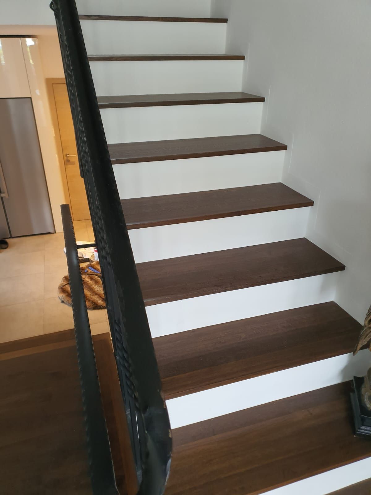 Treppe mit braunen Stufen und weißen Setzstufen, schwarzes Geländer vor einer weißen Wand.