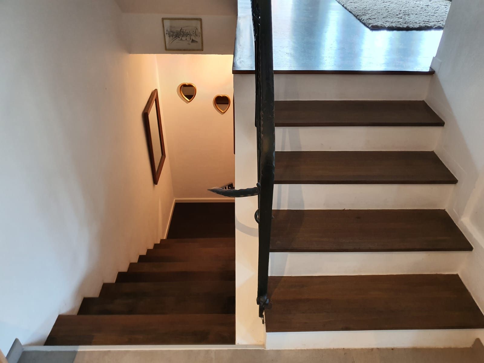 Treppe: Holzstufen führen nach unten, ein schwarzes Geländer verläuft daneben. Licht an den Wänden, Kunstwerke sichtbar.