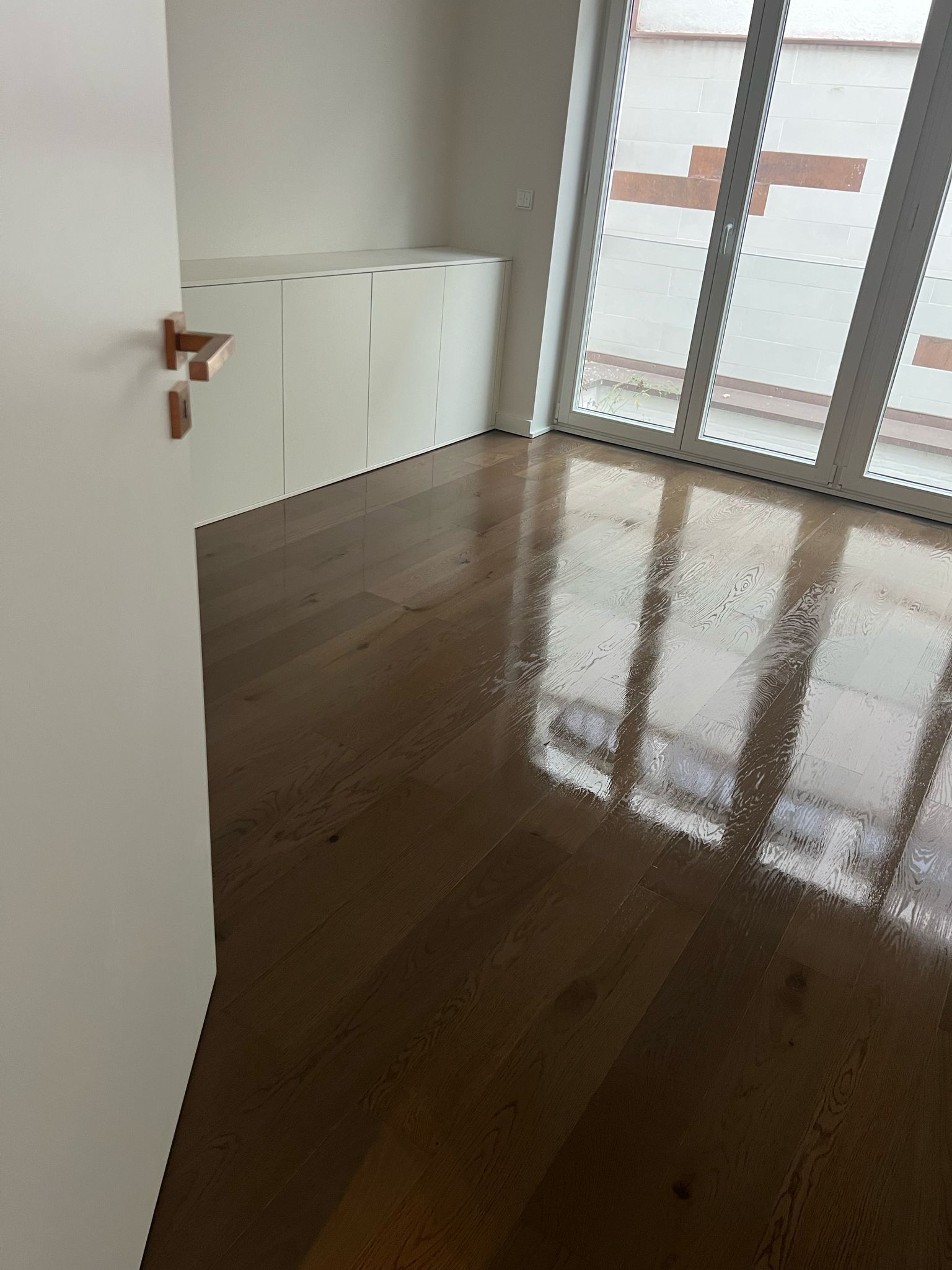 Holzboden in einem Raum mit großem Fenster, das das Tageslicht reflektiert. Weiße Wand und eingebauter Stauraum.