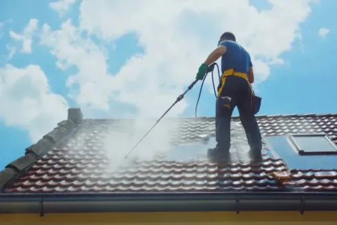 Un trabajador limpia con agua a presión un tejado de tejas rojas bajo un cielo azul.