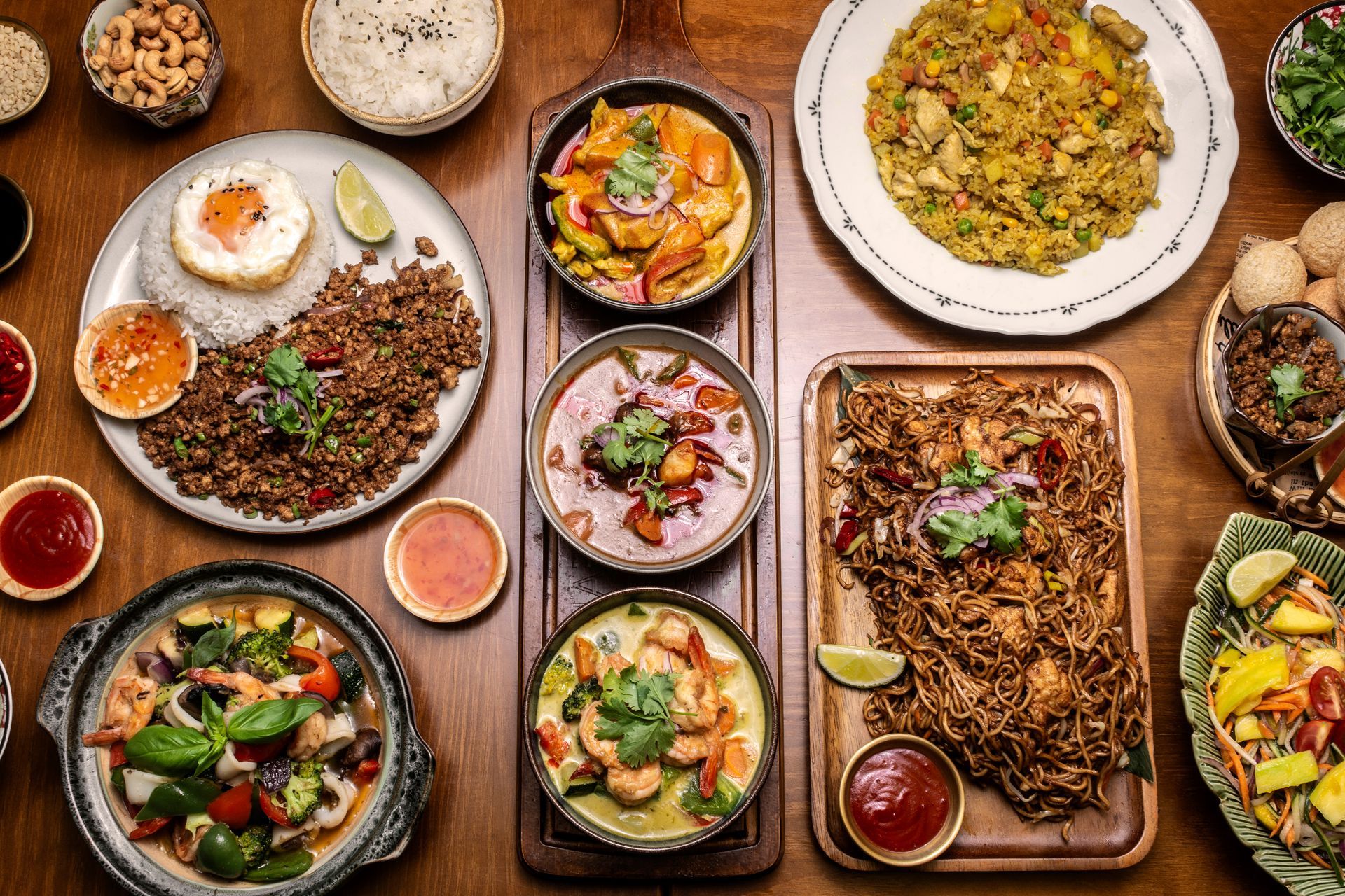 Vue aérienne d'une table en bois chargée de divers plats thaïlandais, notamment des currys, du riz, des nouilles et des sauces.