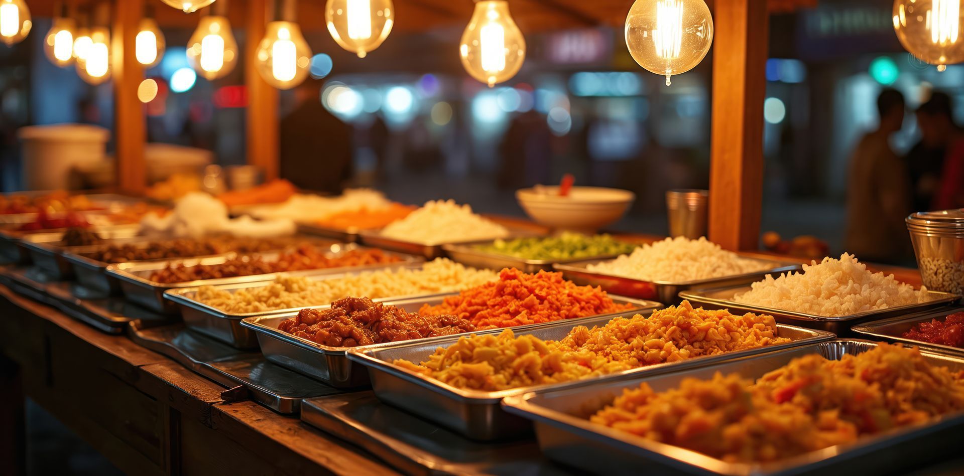 Stand de restauration de rue avec des plateaux de nourriture illuminés sous des ampoules suspendues.