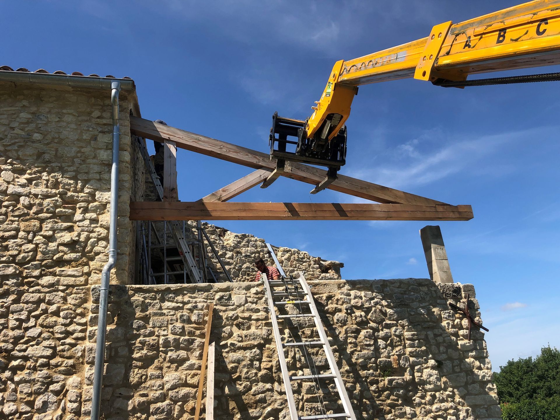 Une pelleteuse soulève une poutre en bois pour la poser sur un bâtiment en pierre en construction, des échelles étant appuyées en dessous.