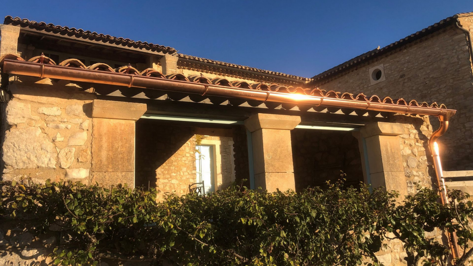 Maison rustique en pierre baignée de soleil, avec toit de tuiles et porche, partiellement cachée par des arbustes.