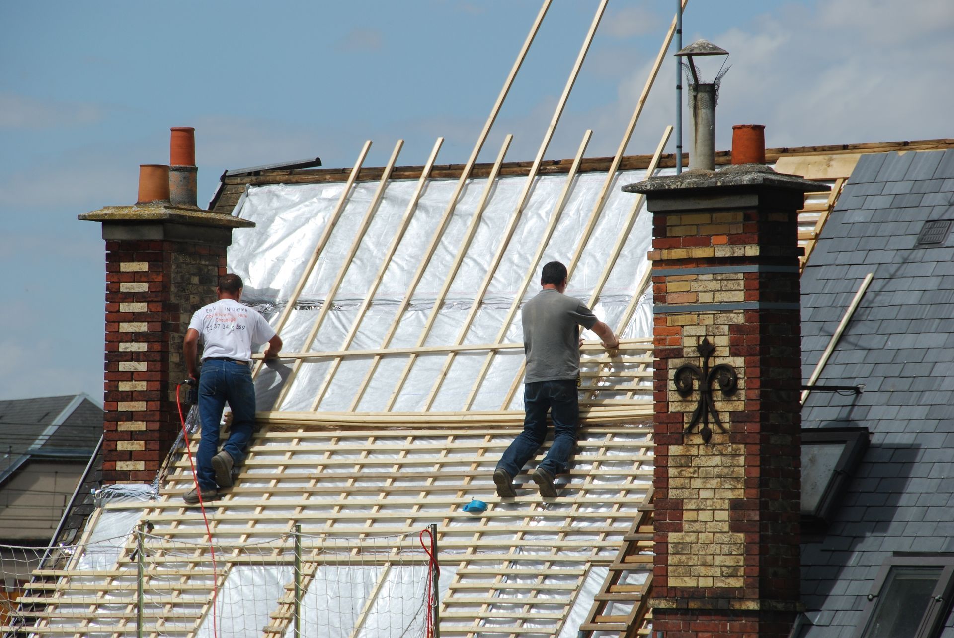 Deux ouvriers installent une charpente de toit sur une maison avec des cheminées en briques.