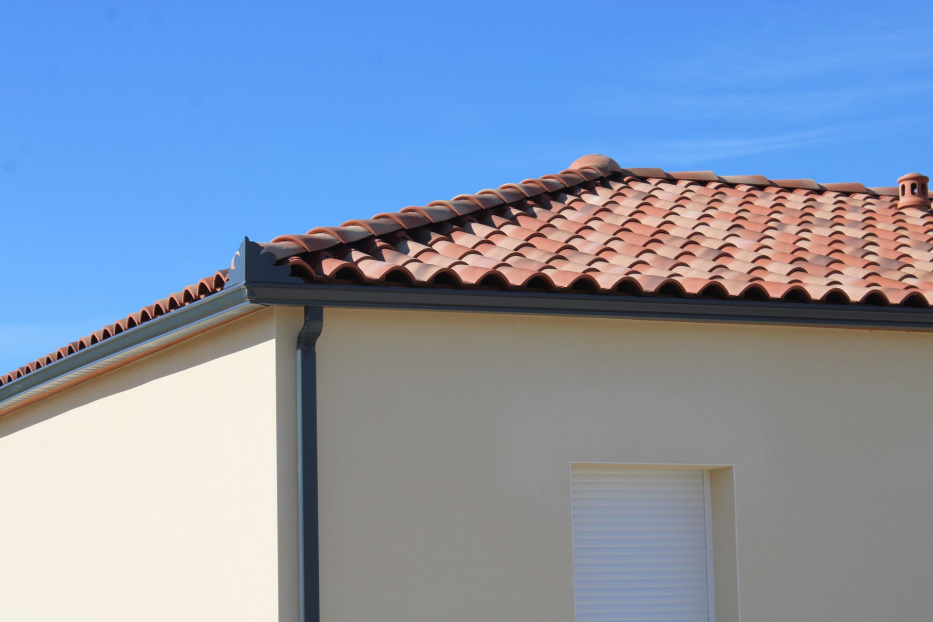 Coin d'une maison couleur crème au toit de tuiles en terre cuite se détachant sur un ciel bleu