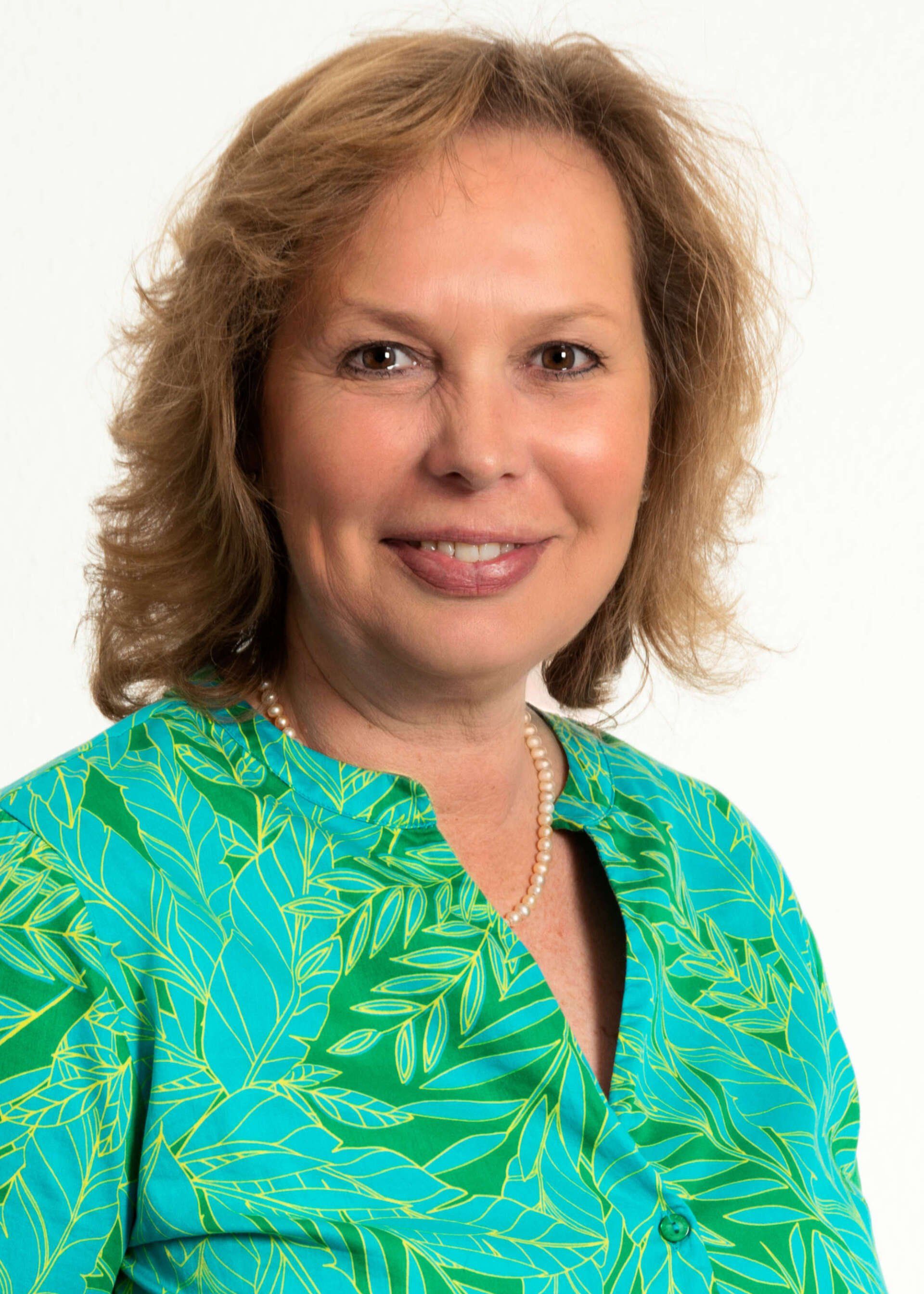 Frau mit kurzem, welligem braunem Haar, trägt ein grün gemustertes Hemd, lächelt und blickt in die Kamera.
