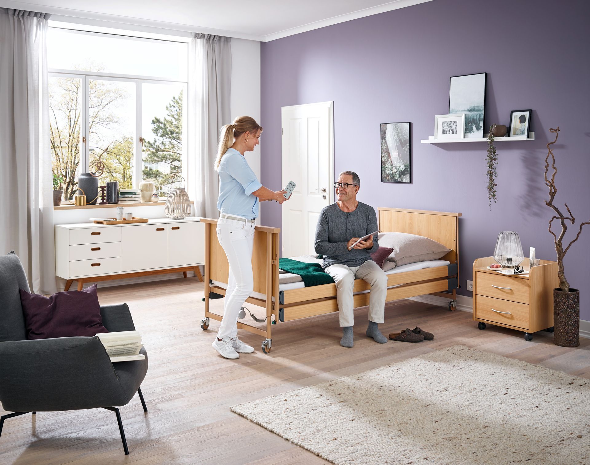 Eine Frau steht neben einem Mann und sitzt in einem Wohnzimmer auf einem Bett.