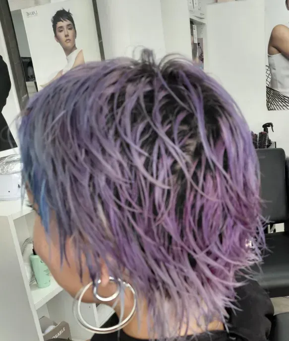 Persona con cabello corto, mojado y de color morado y azul, con grandes aros, en un salón.