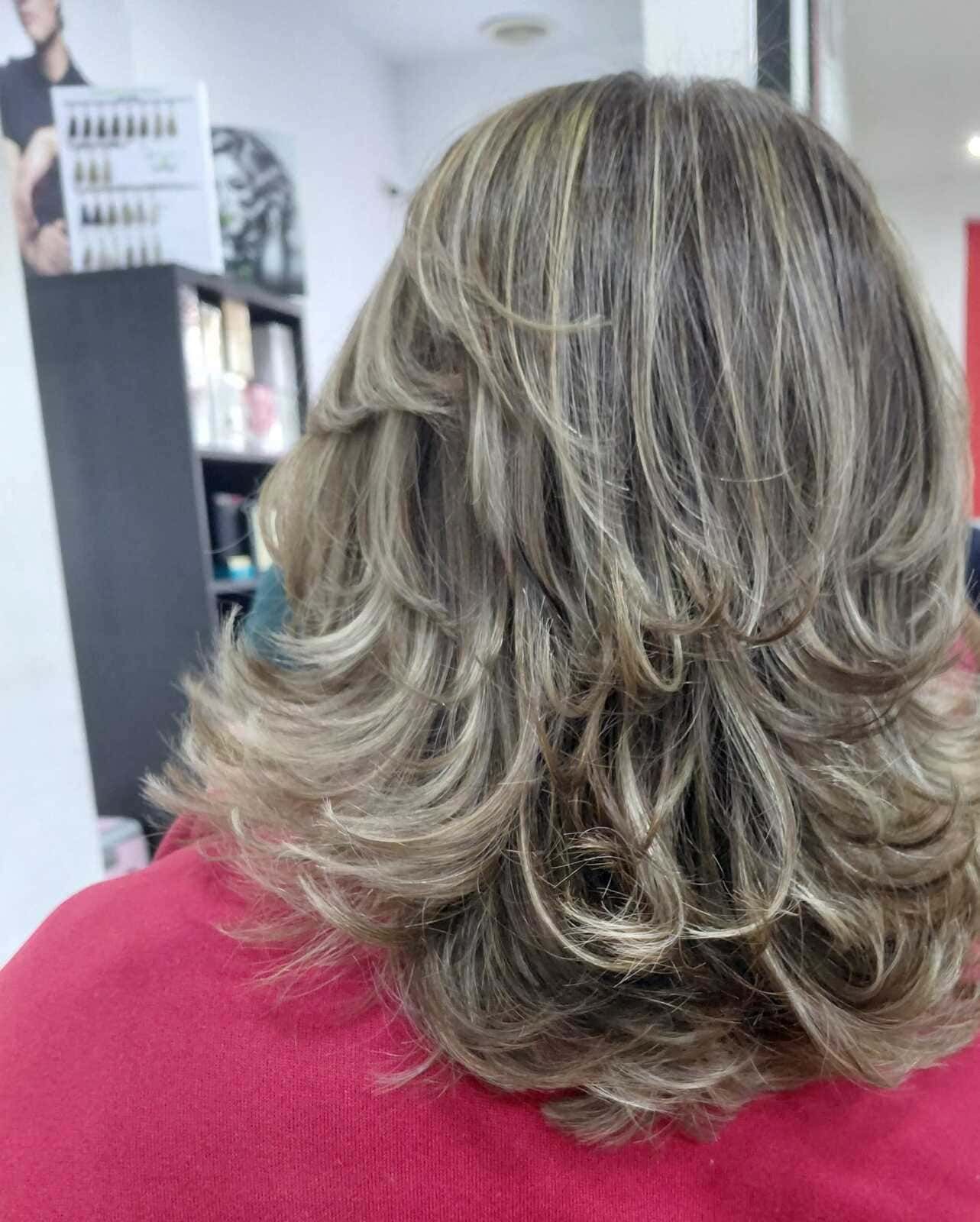 Mujer con cabello en capas, ondulado y con reflejos rubios, vista desde atrás, en una peluquería.